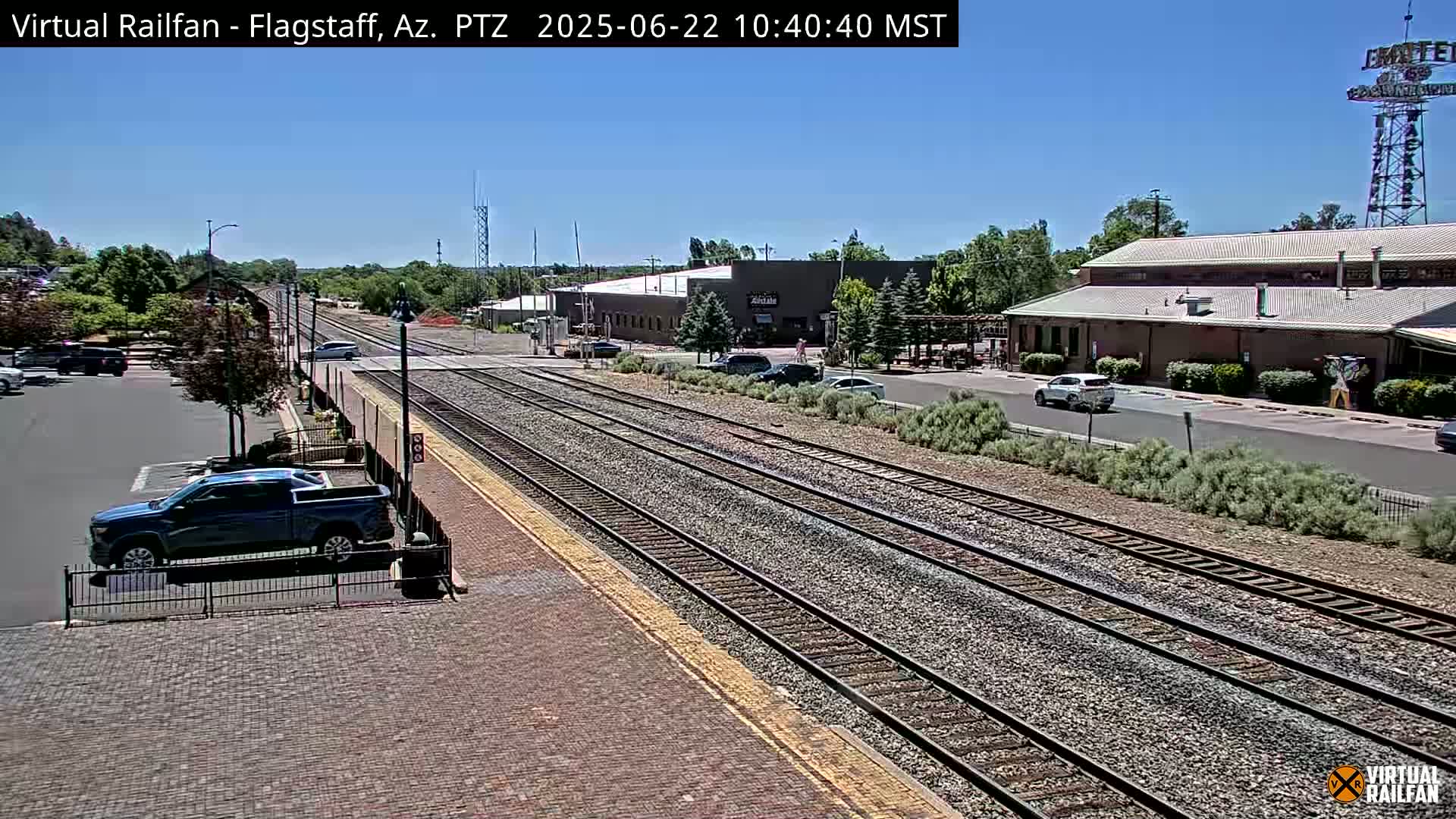 A sunny day reveals a train station with multiple train tracks, a parking lot with cars, and several buildings in the background.