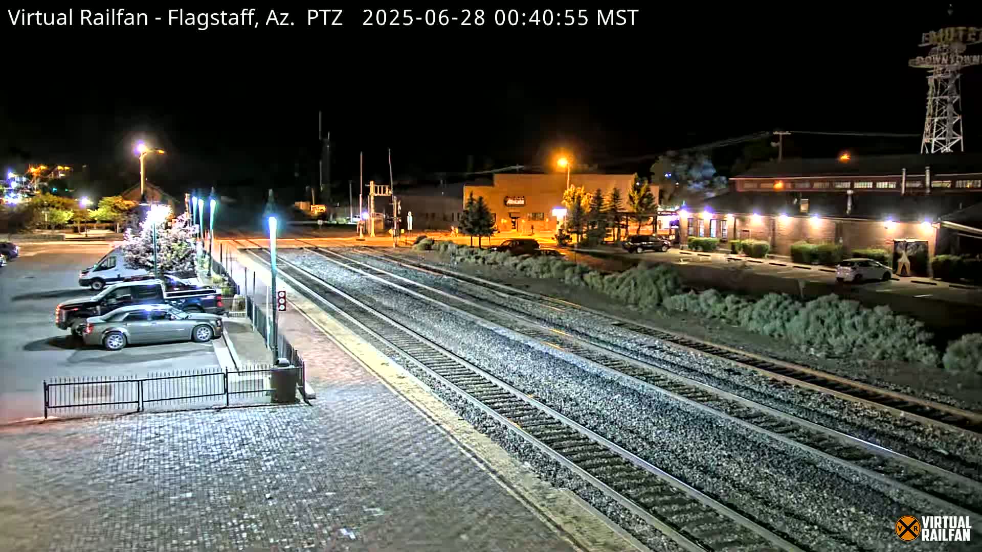 A nighttime view of a train station with multiple train tracks, parked cars in a lot, and a building illuminated by lights.