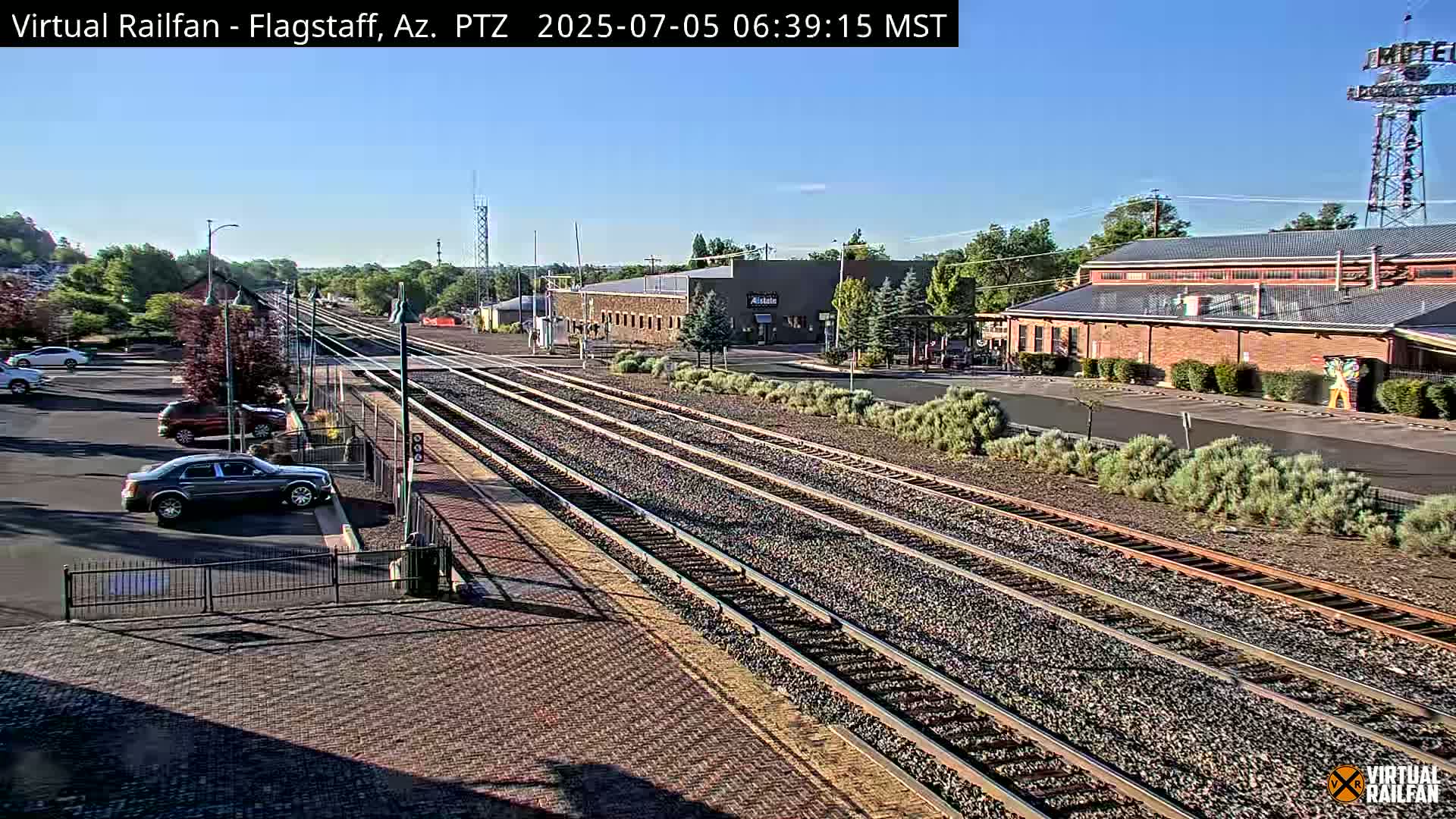 Multiple sets of train tracks extend from a parking lot with several cars, toward a long brick building under a clear, sunny sky.