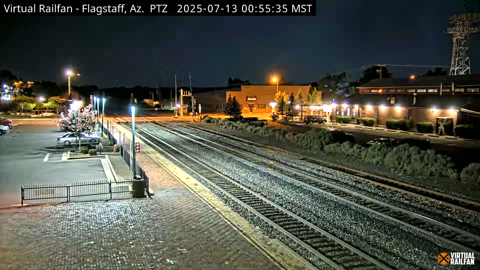 A nighttime view of a train station with a mostly empty parking lot and train tracks extending into the distance under a clear sky.