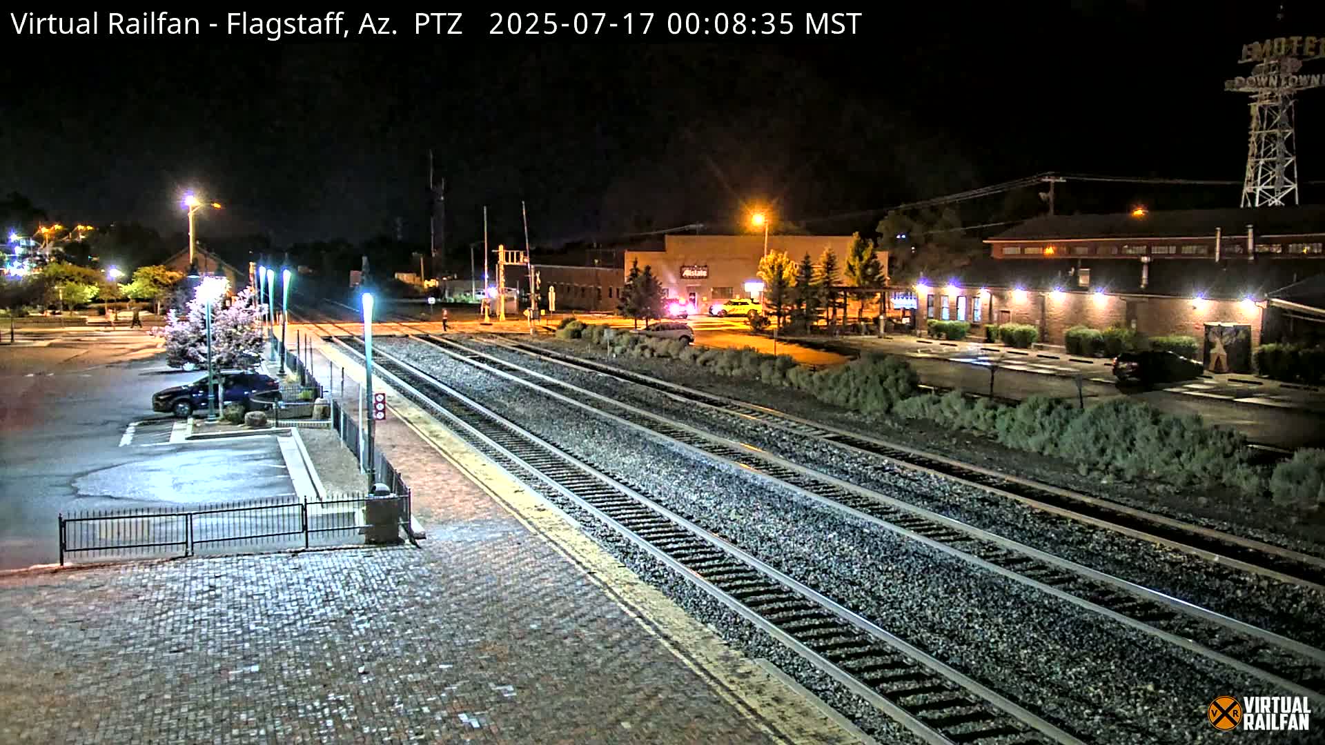 A nighttime view of a train station platform and tracks, with a parking lot and buildings visible in the background under artificial light.