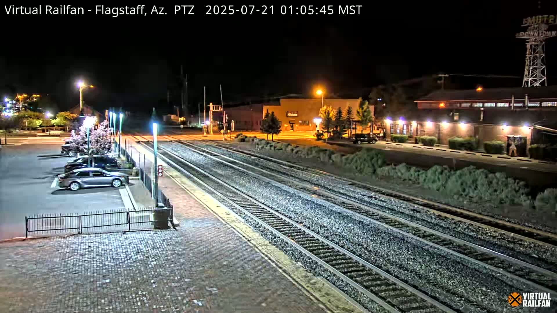 A nighttime view of a train station with several cars parked in a lot adjacent to train tracks and a few buildings in the background.