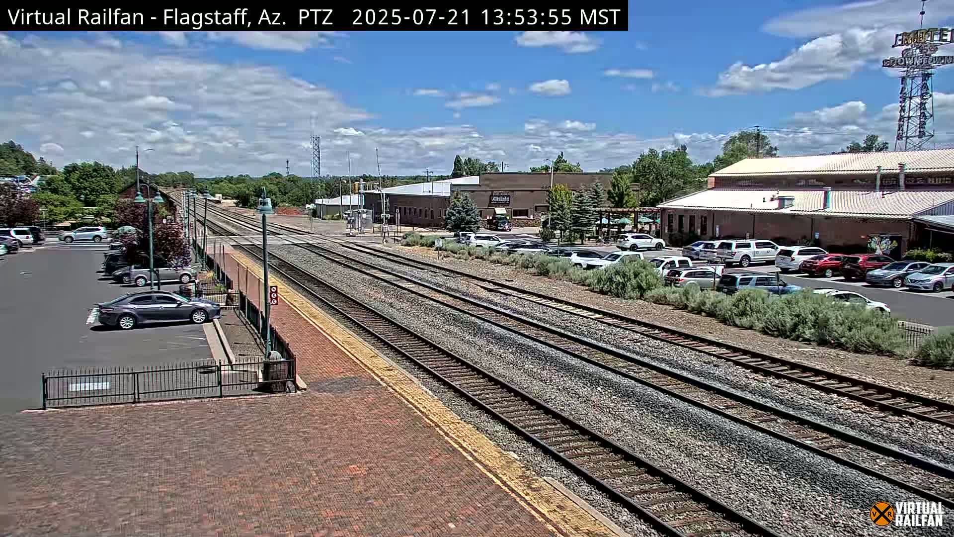 A train track runs through a parking lot with several cars parked near buildings under a partly cloudy sky.