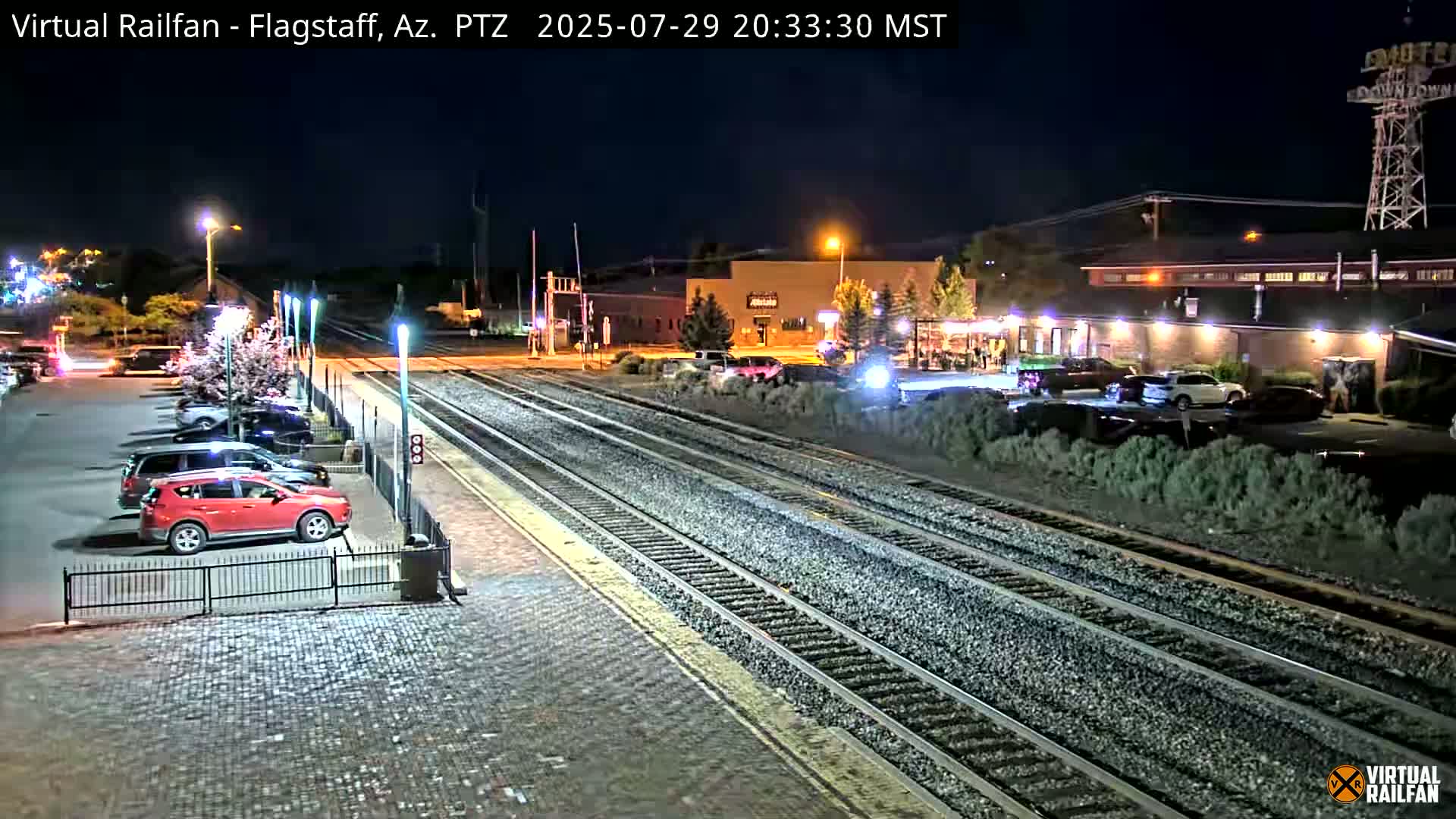 A nighttime view of a train station with multiple train tracks, a parking lot containing a red SUV, and buildings in the background under clear skies.