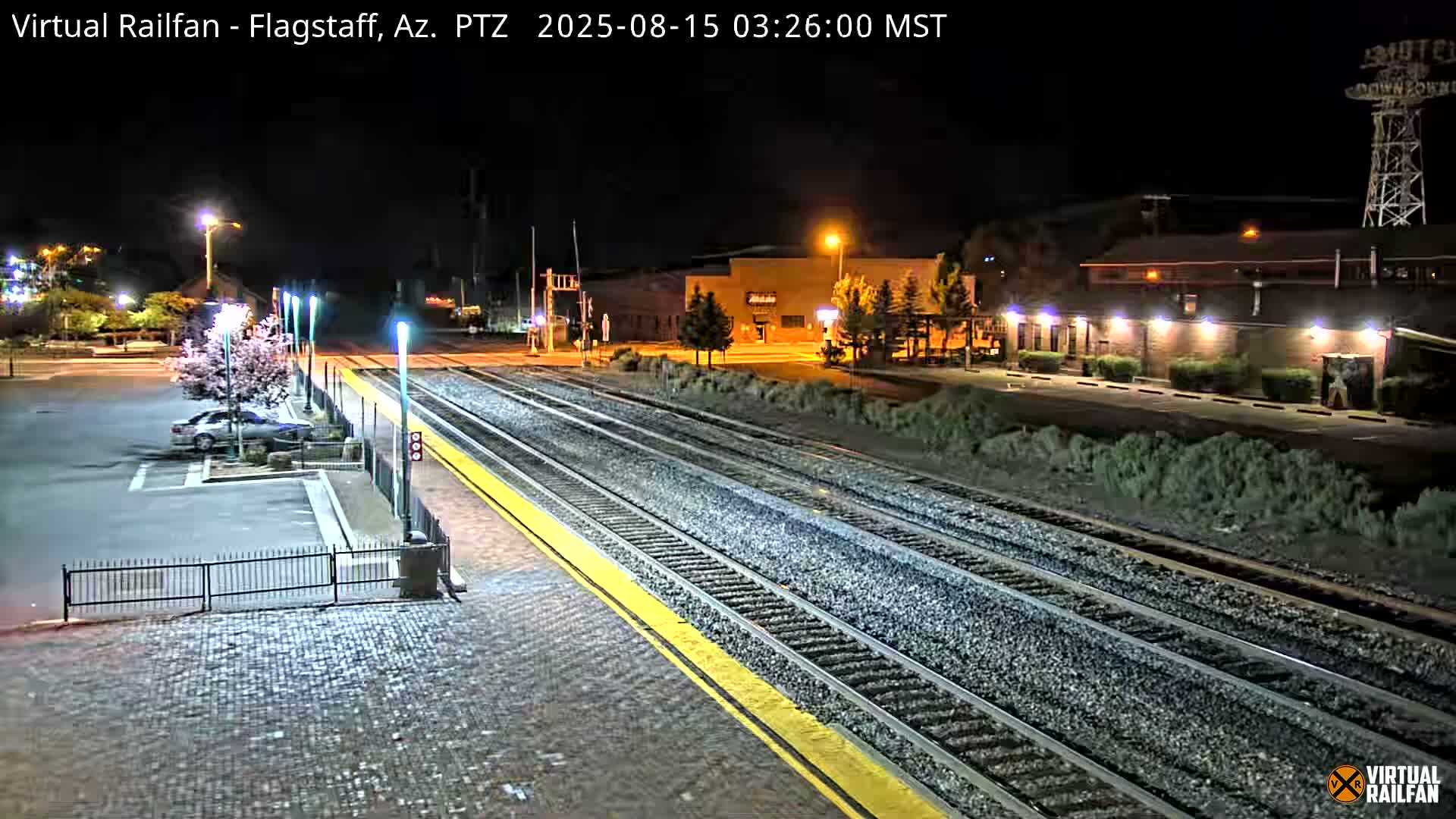 A nighttime view of a train station with multiple train tracks, a parking lot containing a red SUV, and buildings in the background under clear skies.