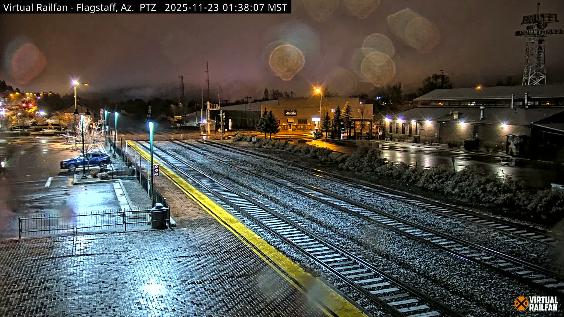 A dark, wet night scene shows glistening train tracks and a cobblestone platform next to a reflective parking lot with a blue SUV, surrounded by lit buildings and mounds of dirty snow.
