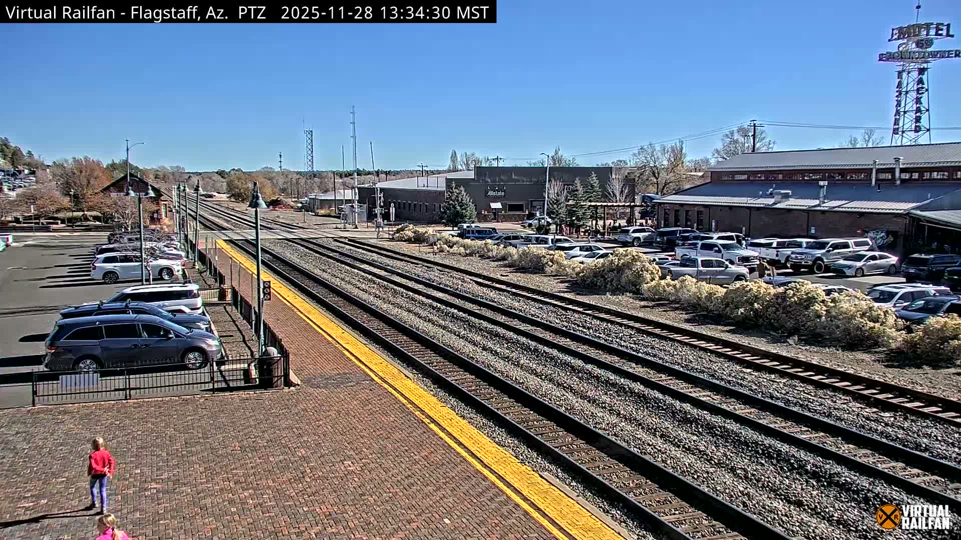 A bright, sunny day illuminates a railroad scene featuring multiple tracks paralleling a brick platform where two children stand, with a busy parking lot and various commercial buildings including one with a vintage motel sign, all under a clear blue sky.