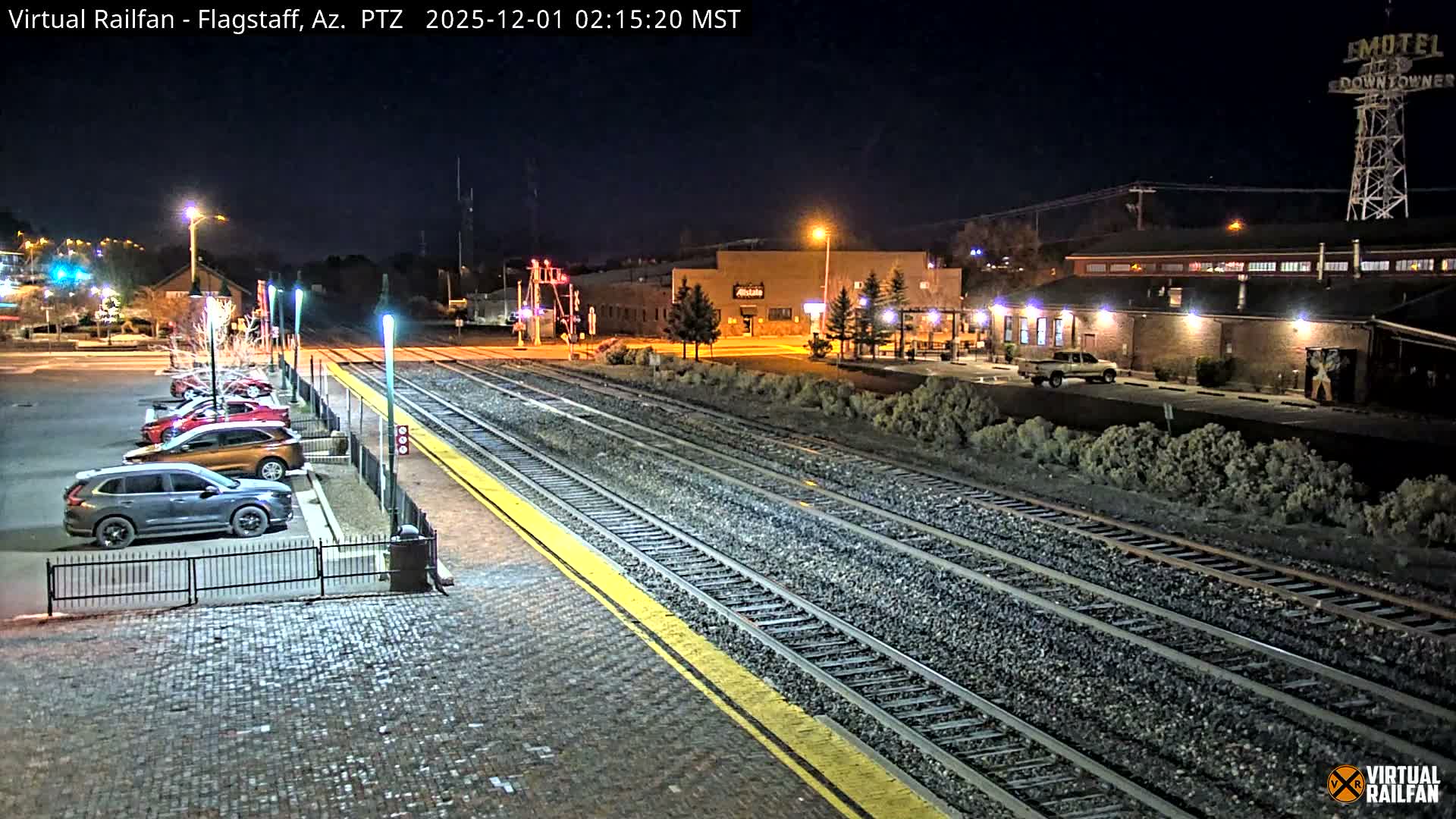The image displays a clear night scene of a train station area featuring multiple tracks with a yellow-lined platform, a cobblestone parking lot with several parked cars, and various illuminated buildings including one with a tall motel sign in the background.