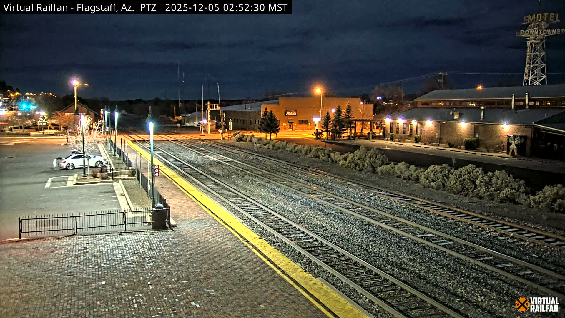 Multiple illuminated train tracks and a yellow-lined platform stretch into the distance, flanked by a cobblestone area, buildings, parked cars, and sparse vegetation under a cloudy night sky.