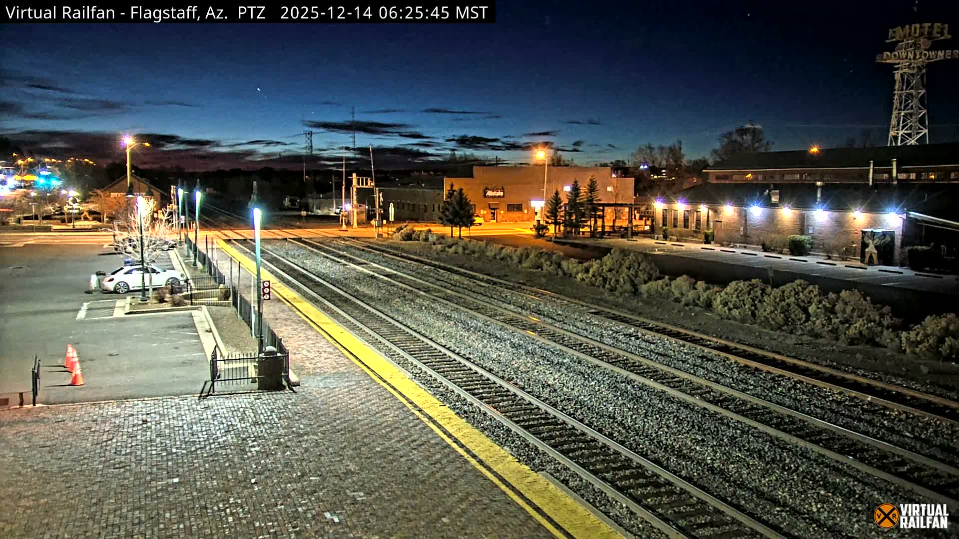Multiple illuminated train tracks and a yellow-lined platform stretch into the distance, flanked by a cobblestone area, buildings, parked cars, and sparse vegetation under a cloudy night sky.