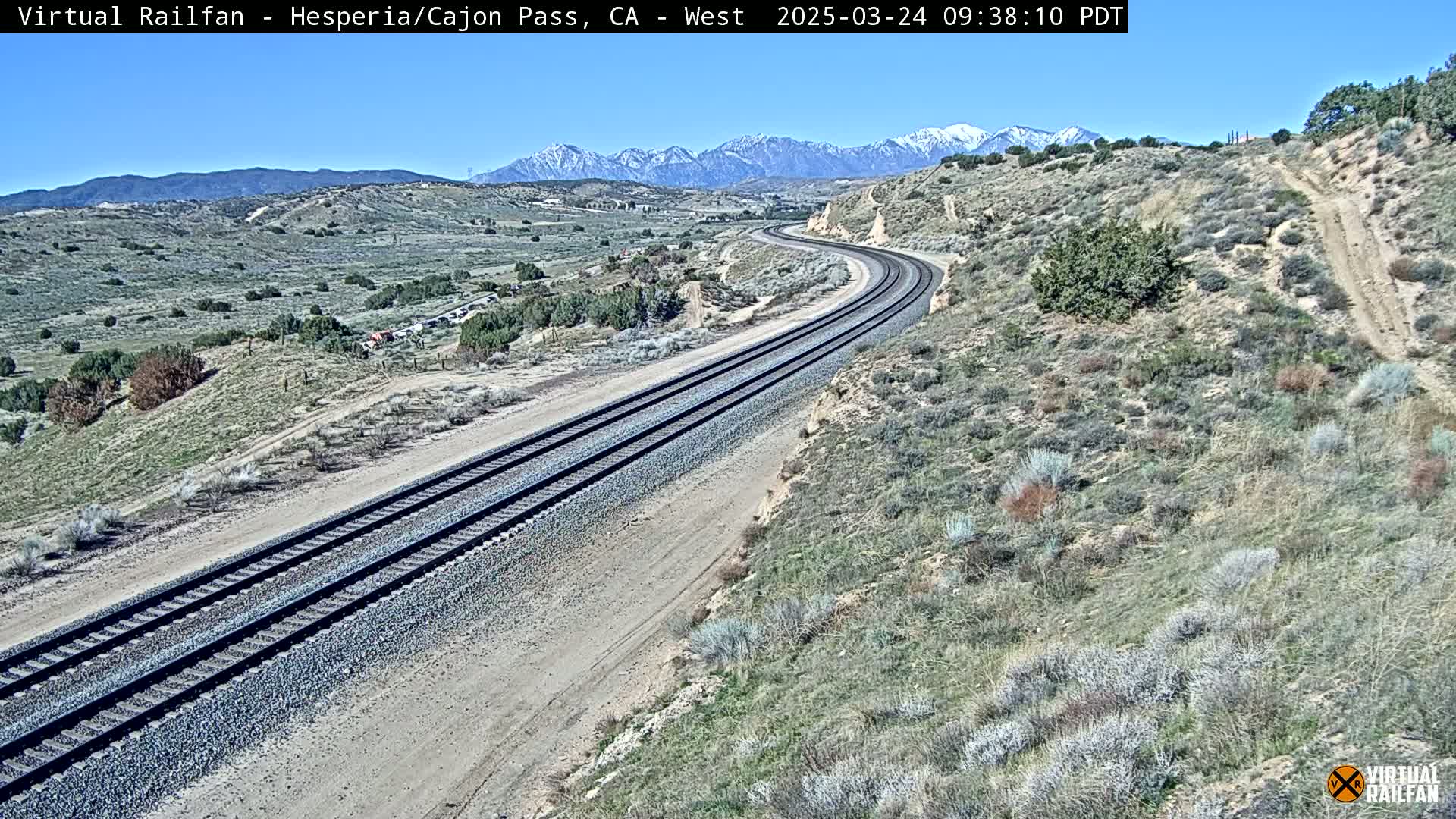 A curving railroad track winds through a dry, desert landscape under a clear blue sky, with snow-capped mountains visible in the distance.