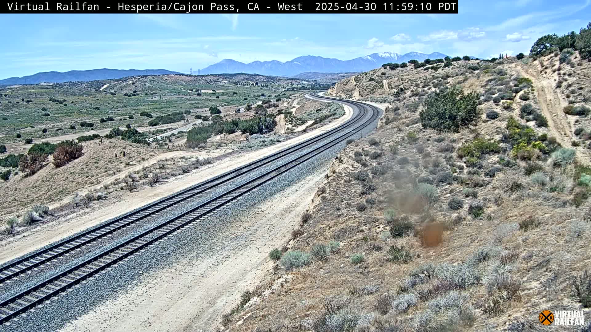 A long, curving railroad track winds through a desert landscape under a clear, sunny sky, with mountains in the distance.