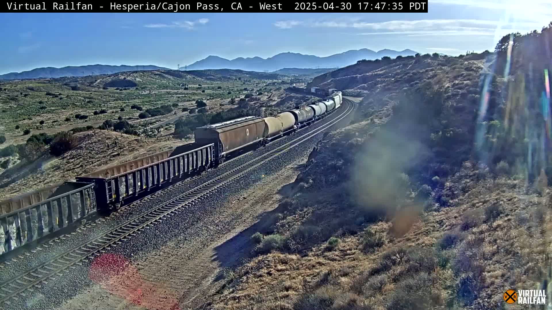 A long freight train travels along railroad tracks through a dry, mountainous landscape under a clear, sunny sky.