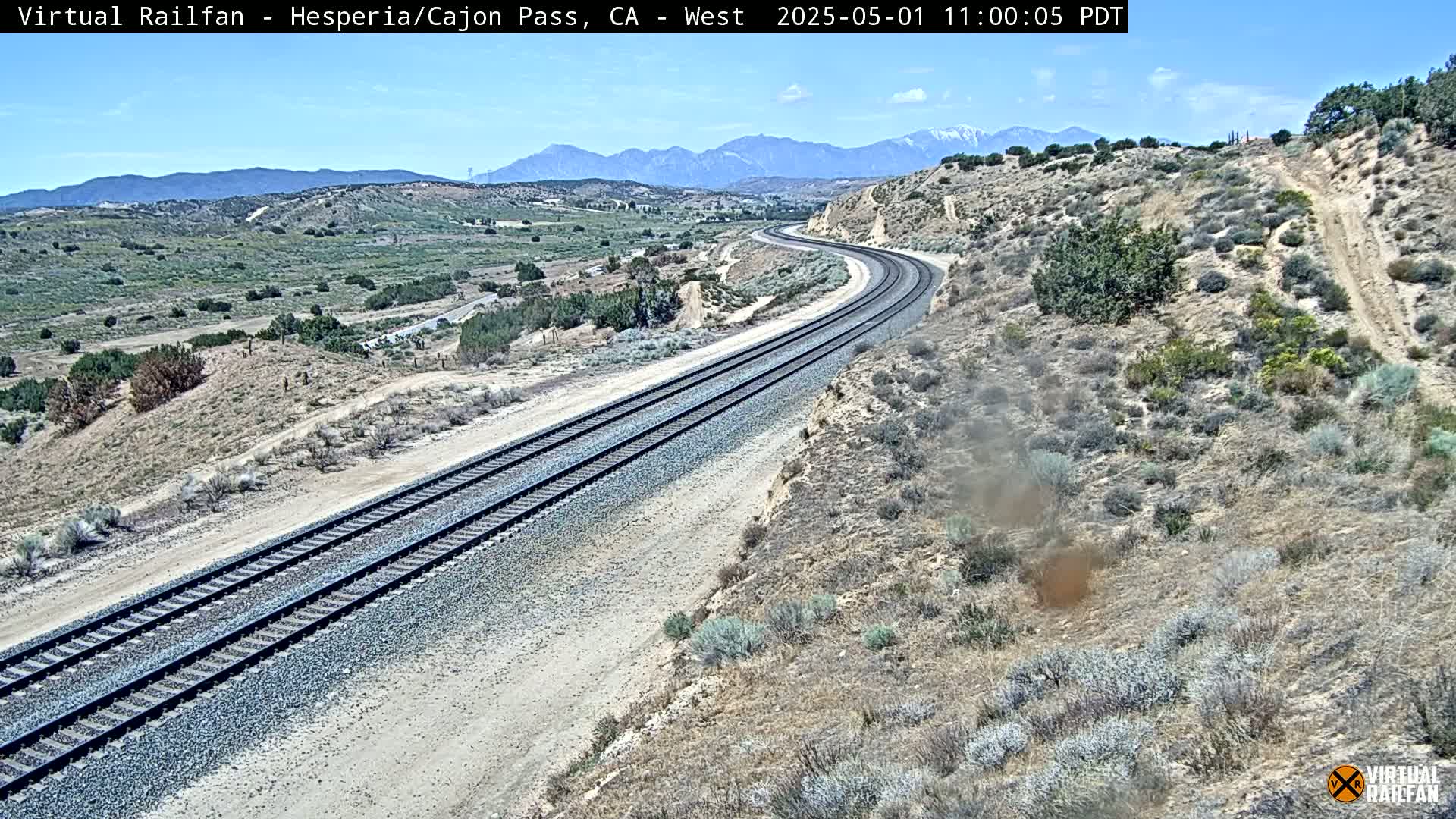 A curving railroad track stretches across a desert landscape under a clear sky, with mountains visible in the distance.