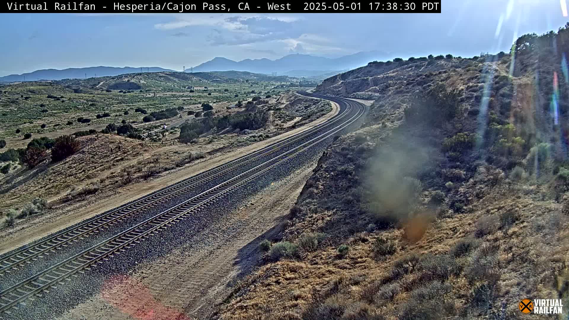A curving railroad track stretches across a dry, hilly landscape under a mostly clear sky.