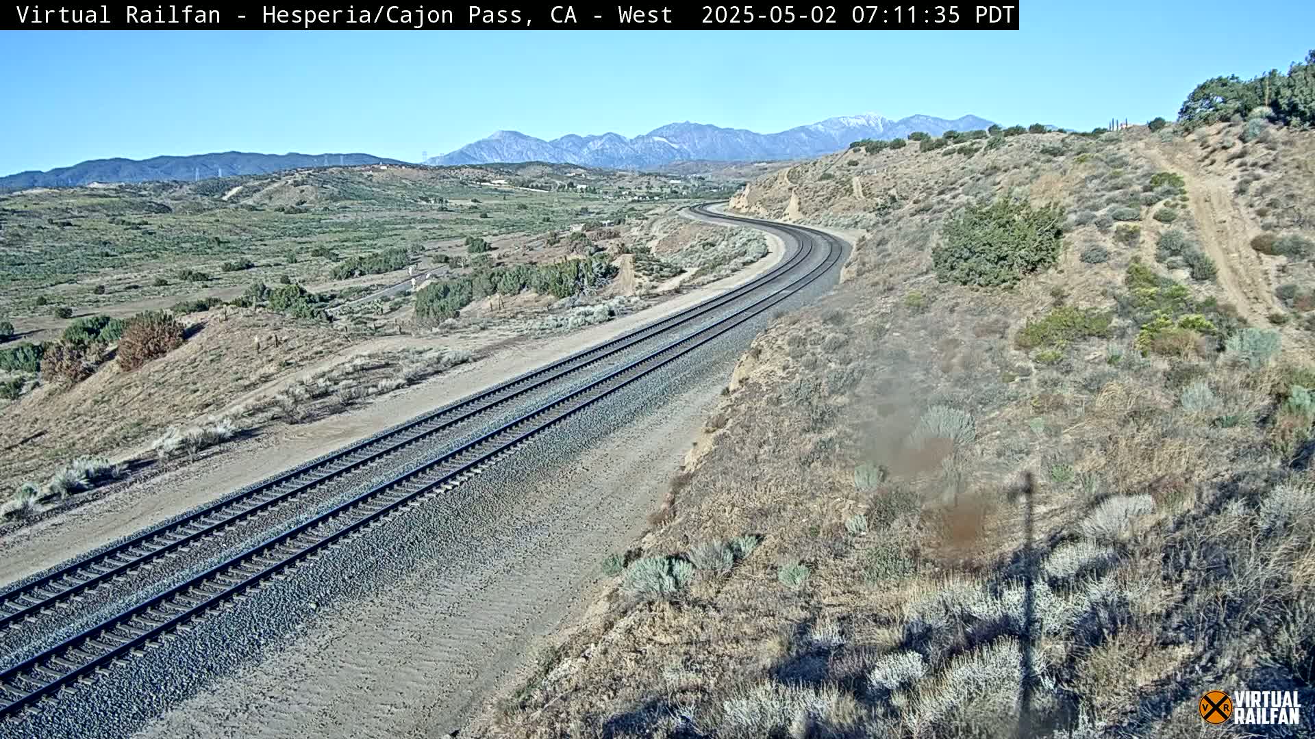 A curving railway track stretches across a dry, desert-like landscape under a clear, sunny sky, with mountains visible in the distance.