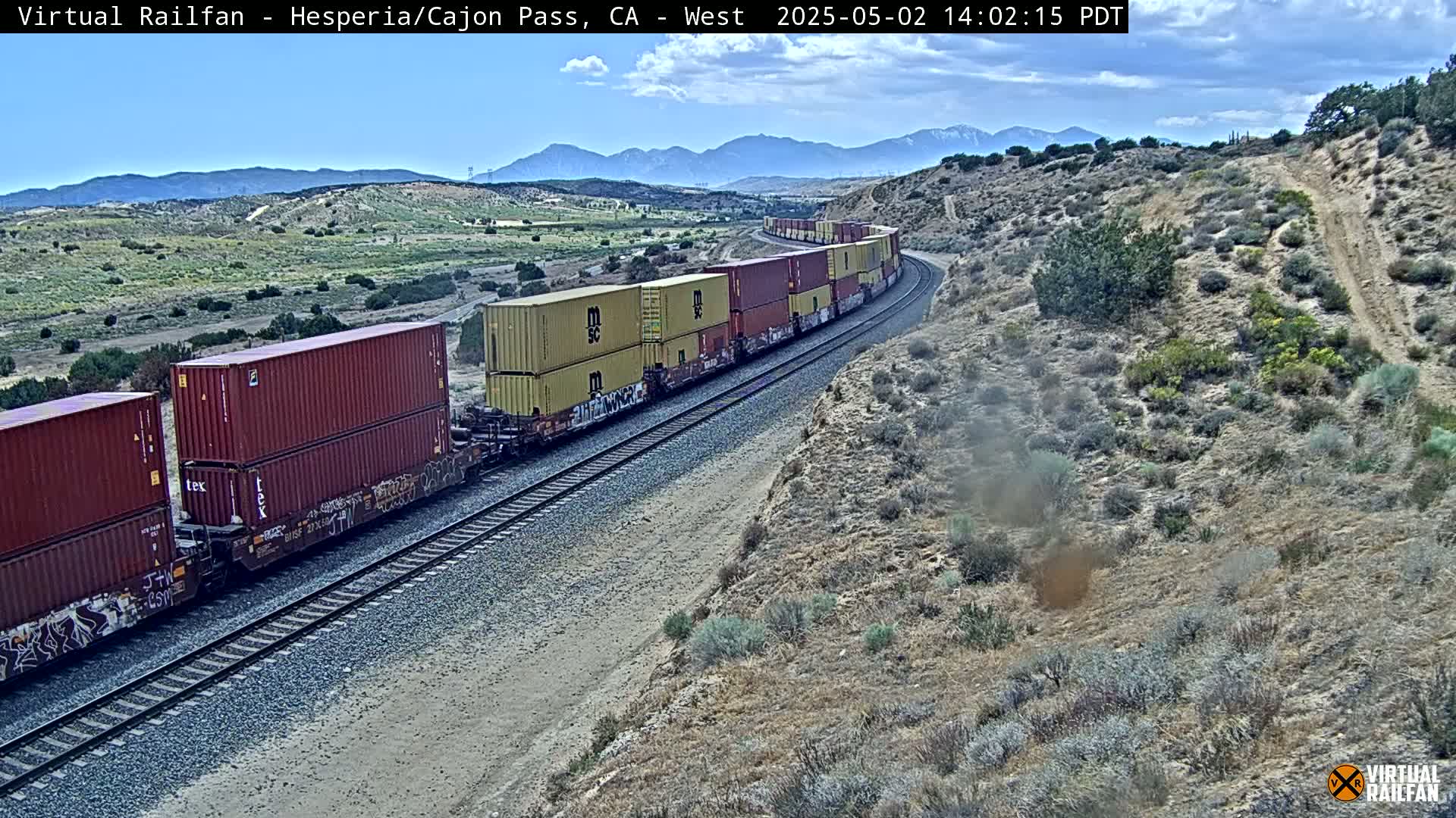 A long freight train carrying stacked shipping containers curves along a railroad track through a dry, hilly landscape under a partly cloudy sky.