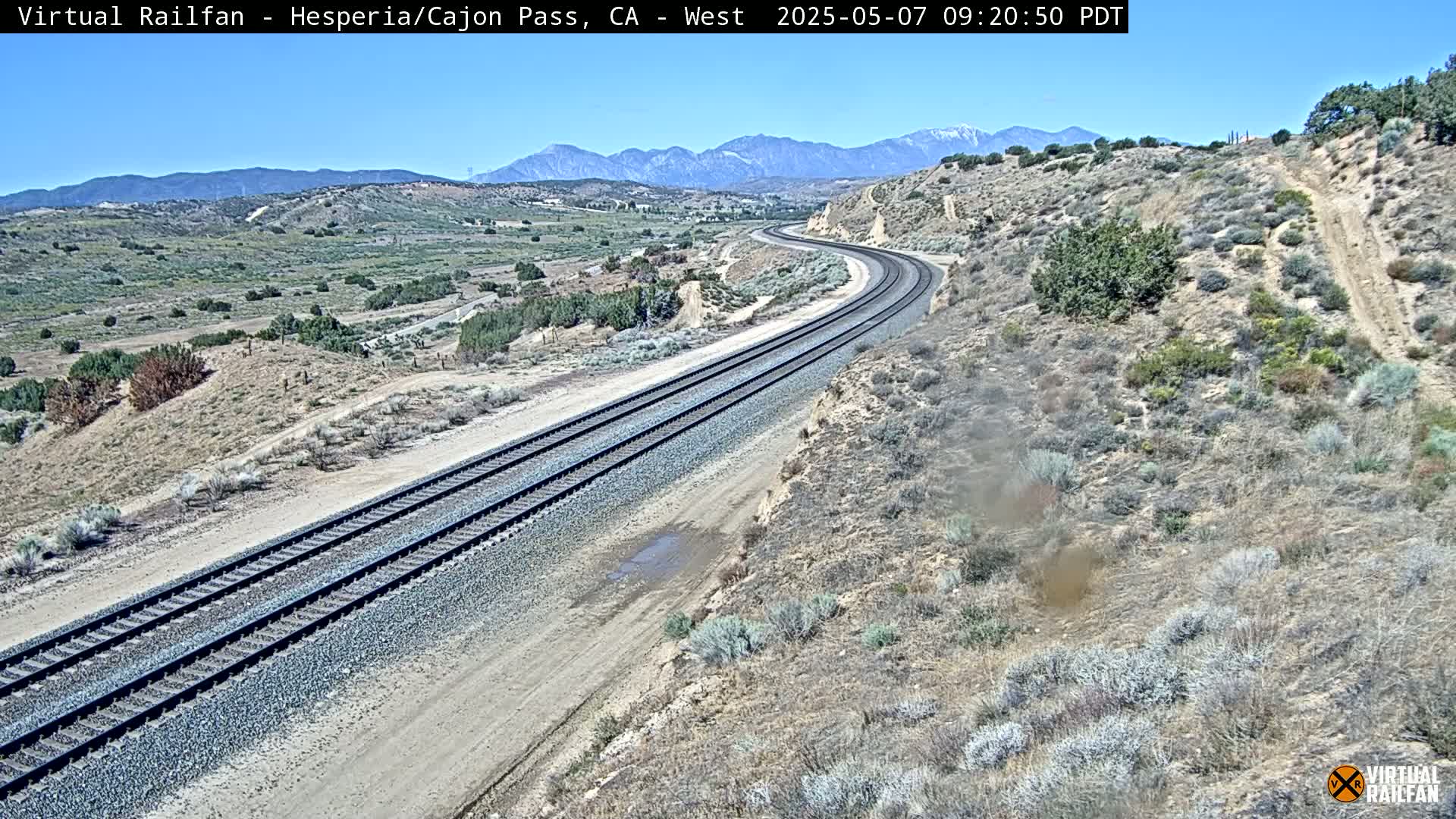 A high-angle view shows a curving train track winding through a dry, desert-like landscape under a clear blue sky, with mountains in the background.
