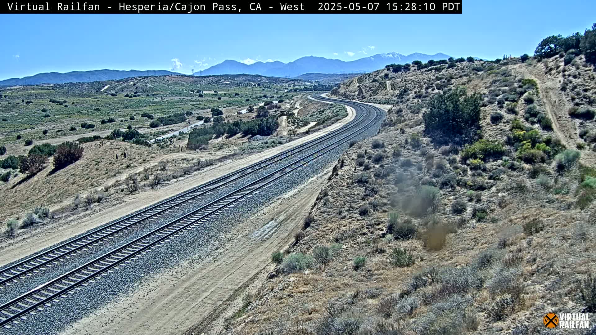 A long stretch of train tracks curves through a dry, desert-like landscape under a clear blue sky, with mountains visible in the distance.
