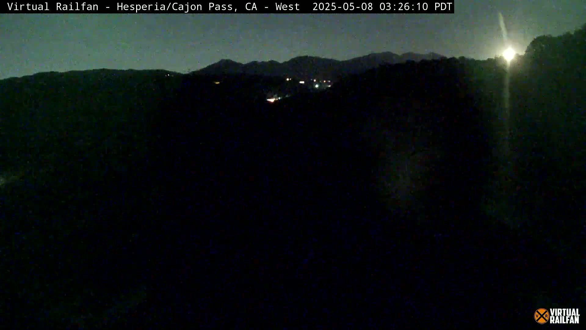 A nighttime view of dark hills and a few distant lights under a mostly clear sky.