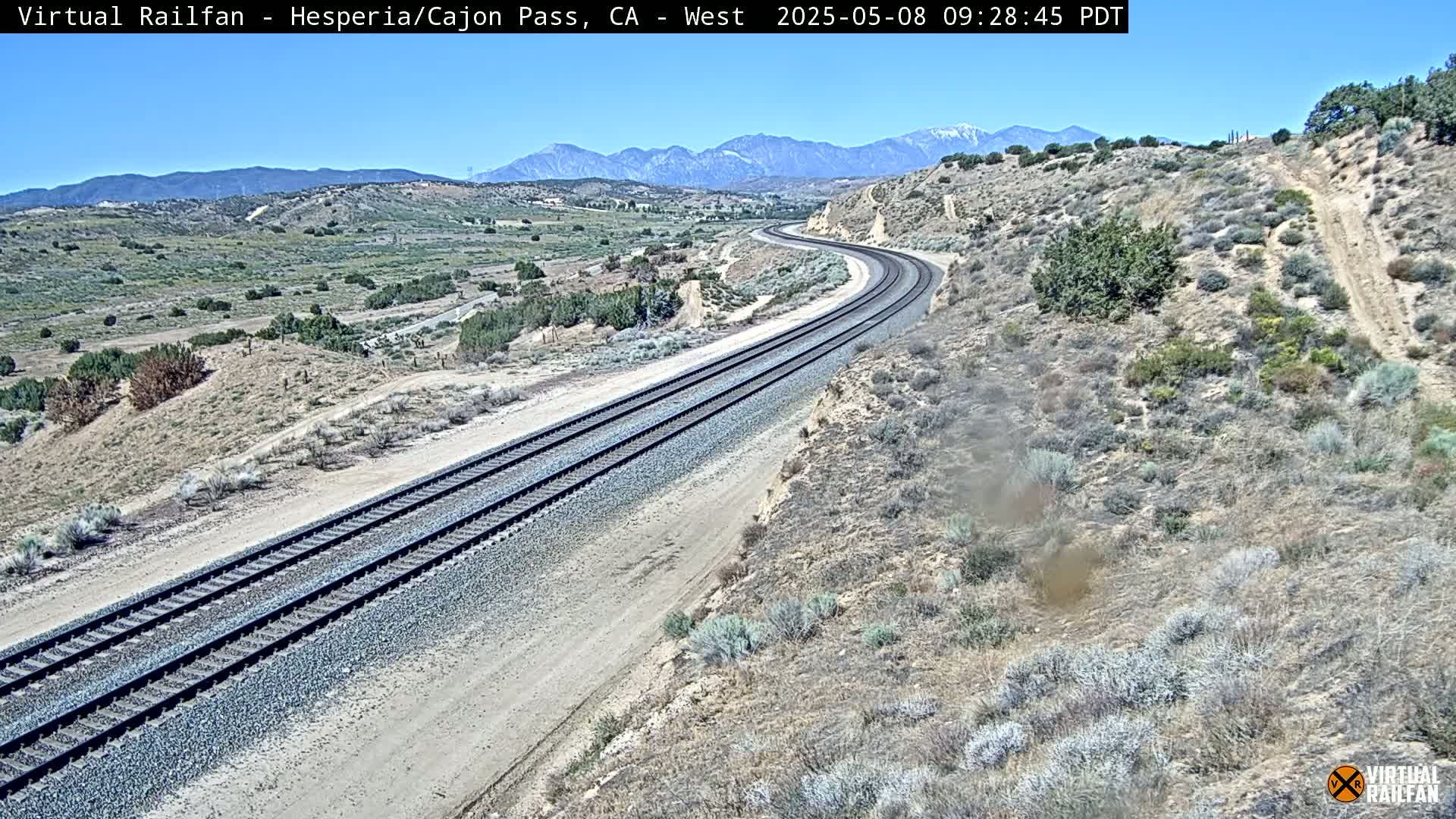 A pair of train tracks curves gently through a dry, desert landscape under a clear, sunny sky, with mountains visible in the distance.