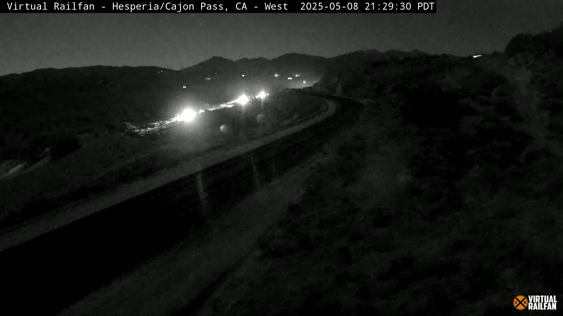 A nighttime, low-light view of a highway curving through a mountainous area, with the headlights of several vehicles visible in the distance.