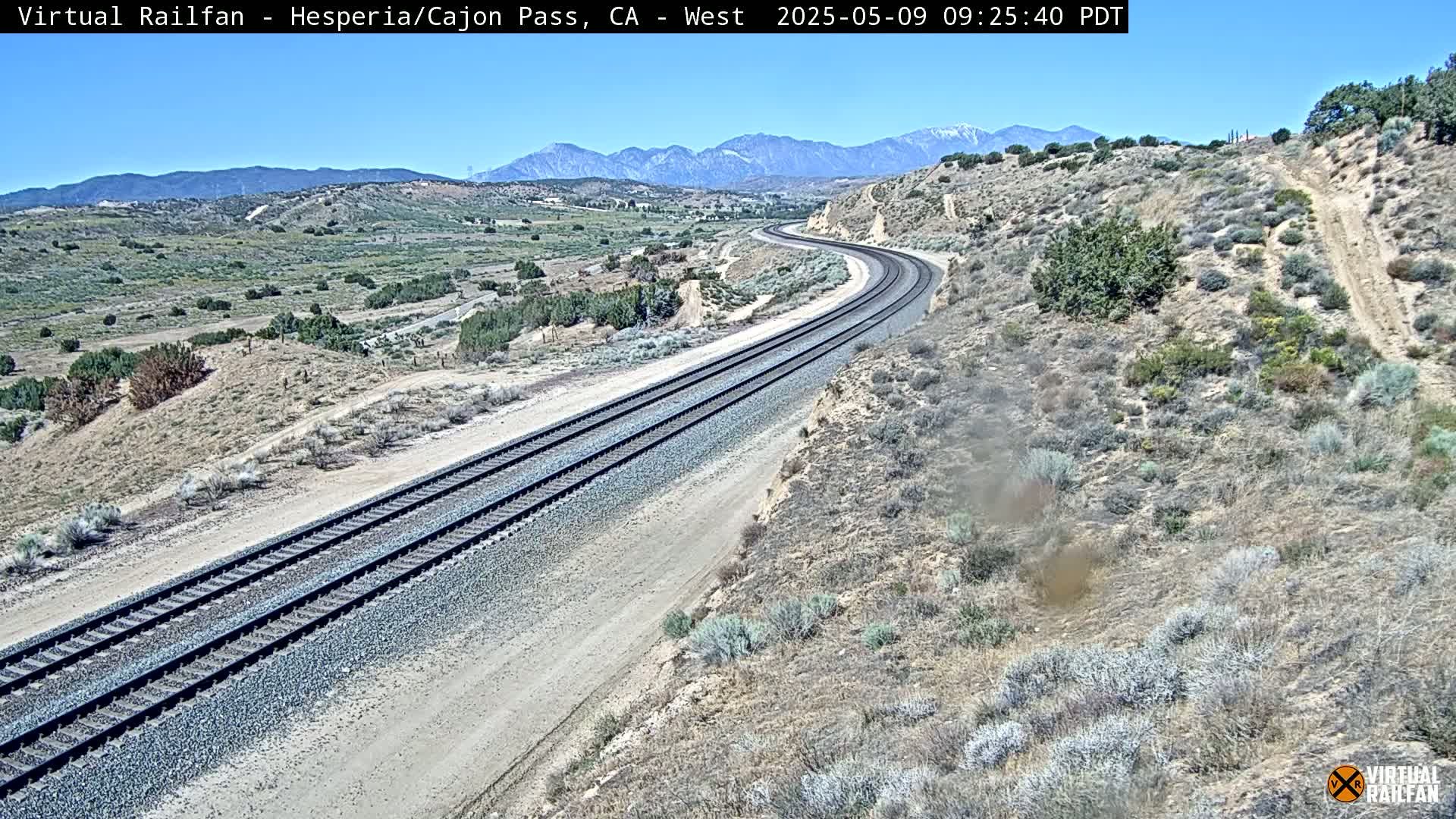 A long stretch of train tracks curves gently through a dry, scrub-covered landscape under a clear blue sky, with mountains in the distance.