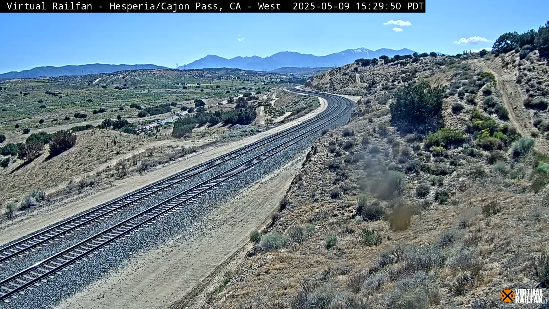 A high-angle view shows a double set of train tracks curving through a dry, desert-like landscape under a clear, sunny sky, with mountains in the distance.