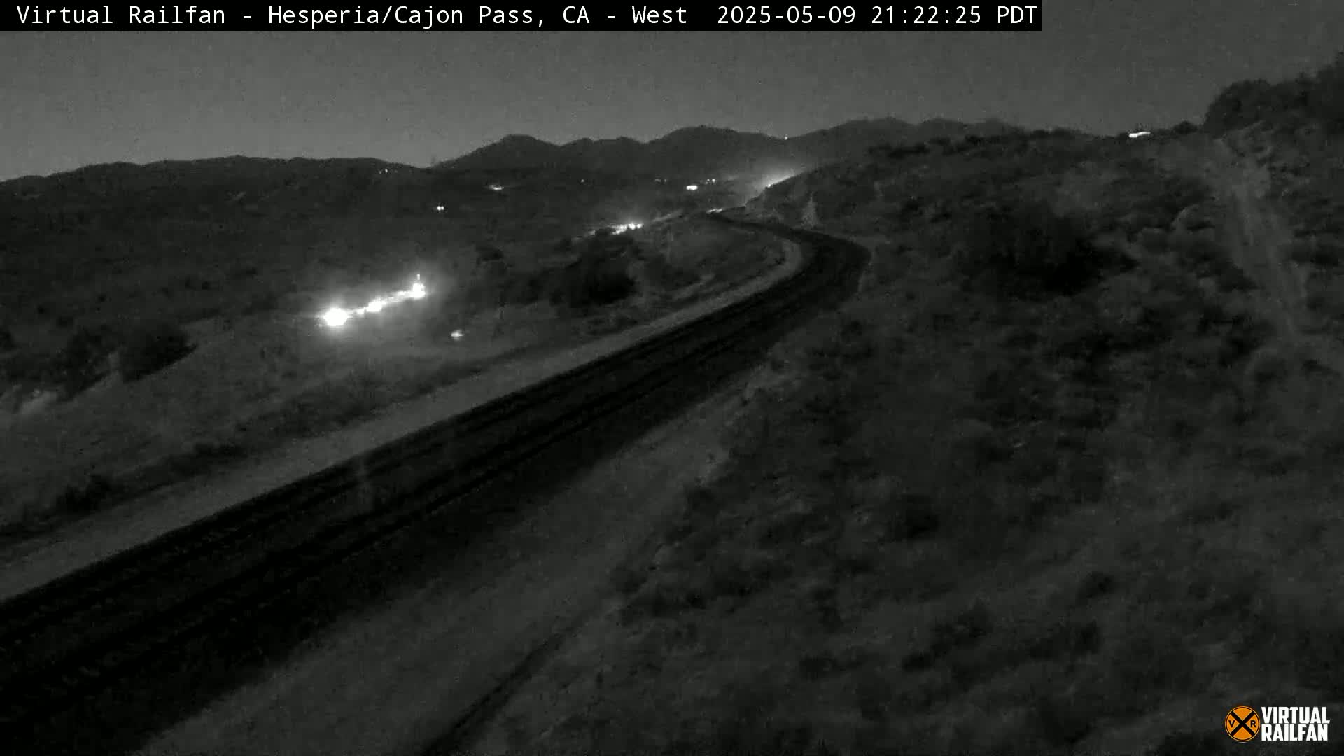 A nighttime, low-light view of a curving roadway and train tracks cutting through a sparsely vegetated landscape under a dark sky.