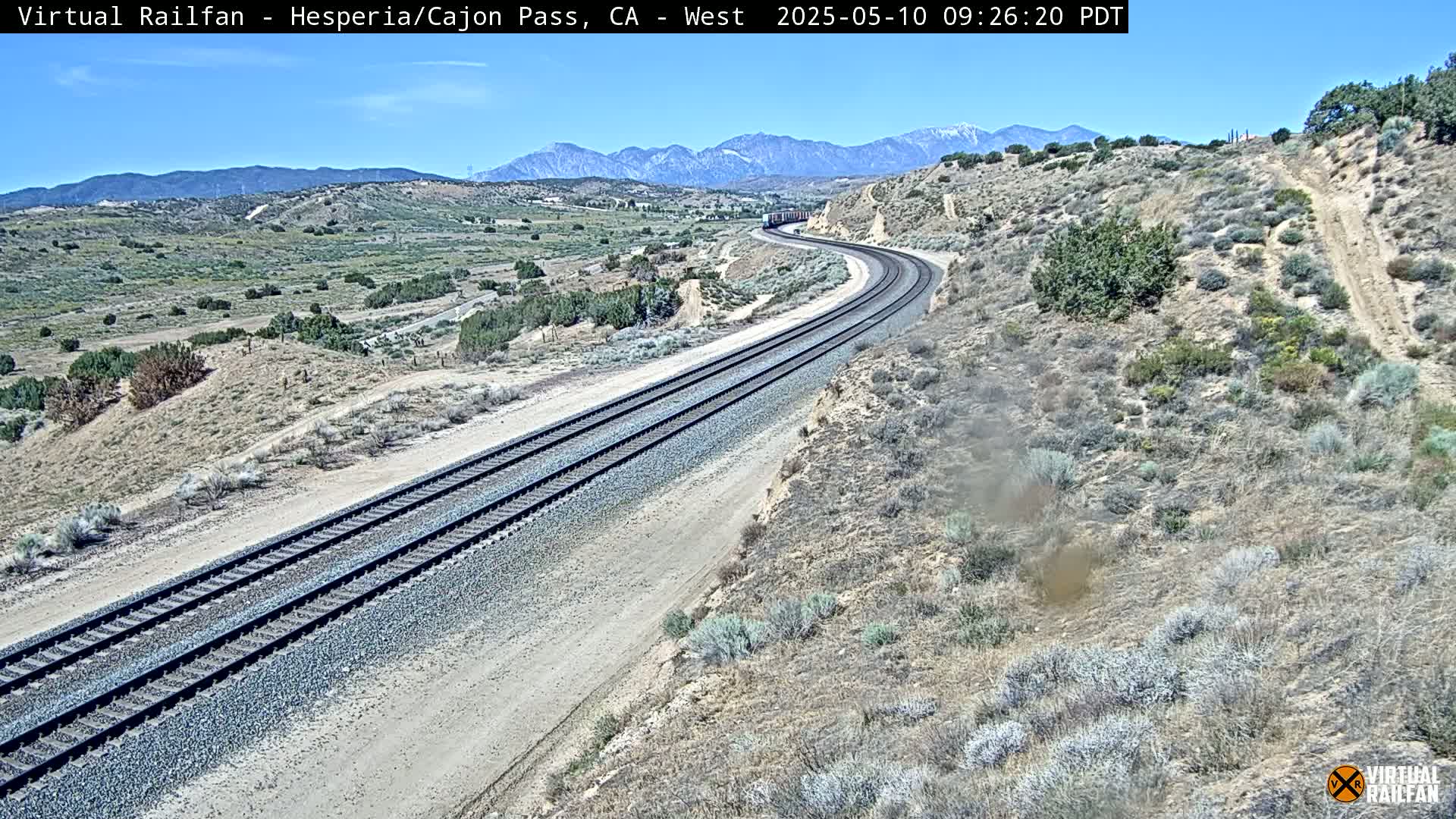 A long freight train curves along double railroad tracks through a desert landscape under a clear blue sky, mountains visible in the distance.