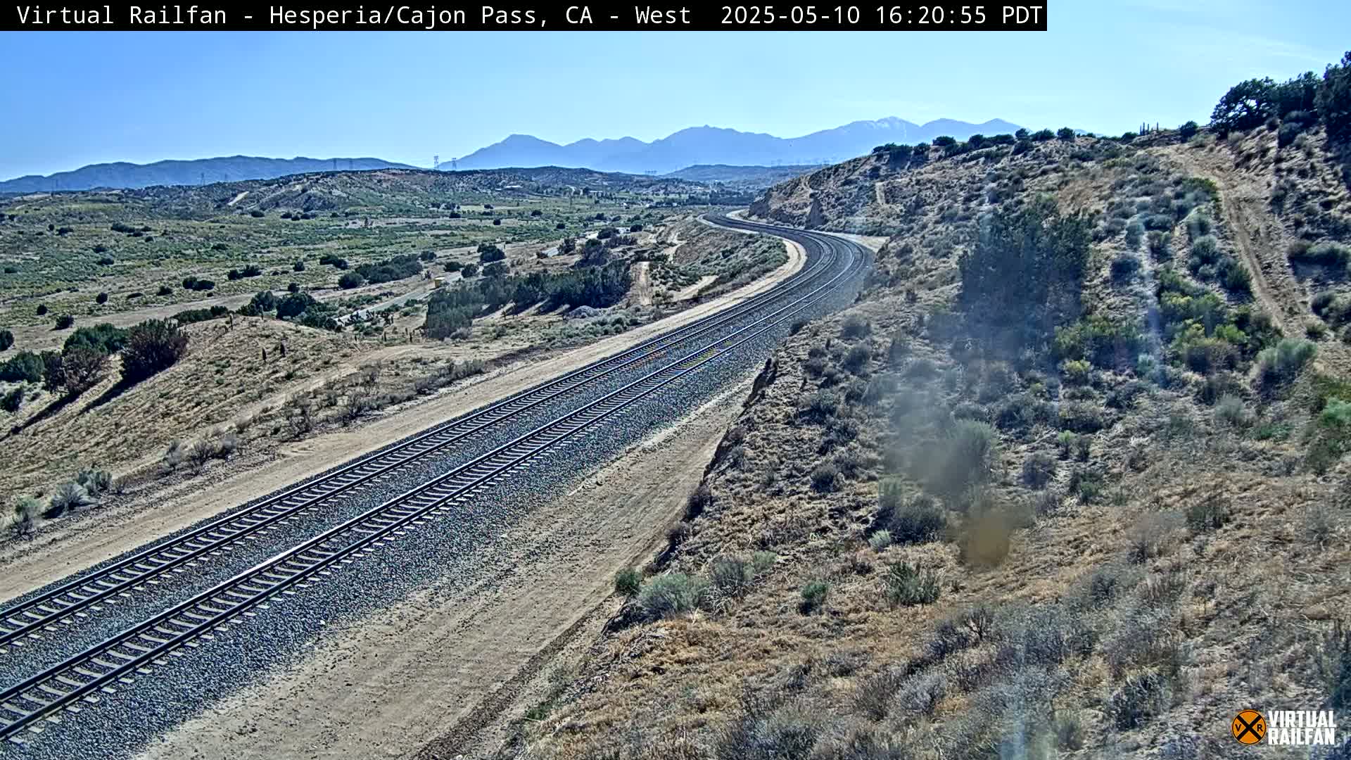 A high-angle, long shot of a curving double railway track traversing a dry, desert-like landscape under a clear, sunny sky, with mountains visible in the distance.