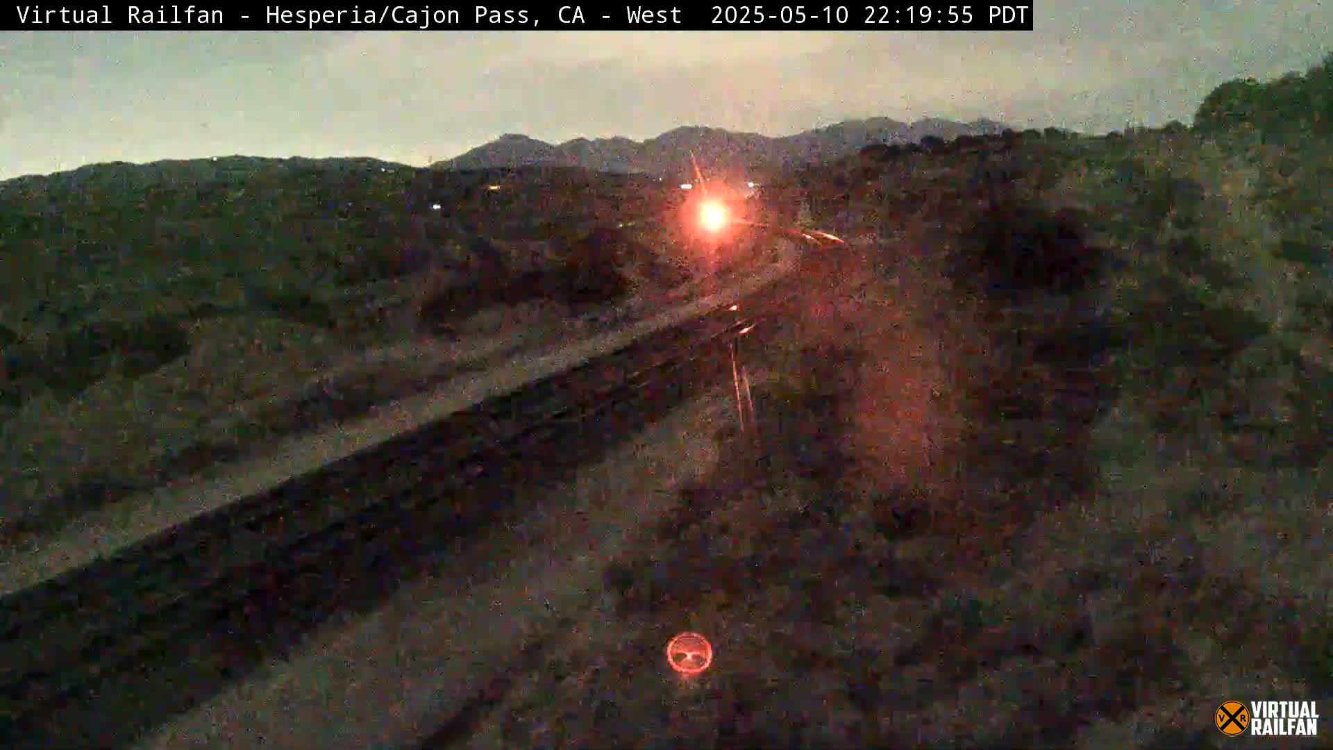 A nighttime, low-light view of a train traveling along railroad tracks in a desert-like landscape.