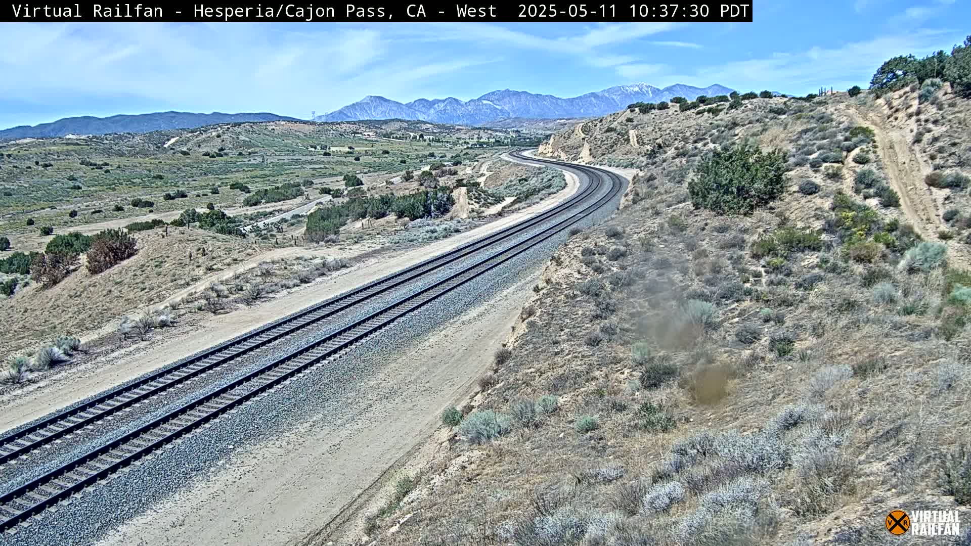A high-angle view shows a curving railway track stretching towards mountains under a mostly clear sky.