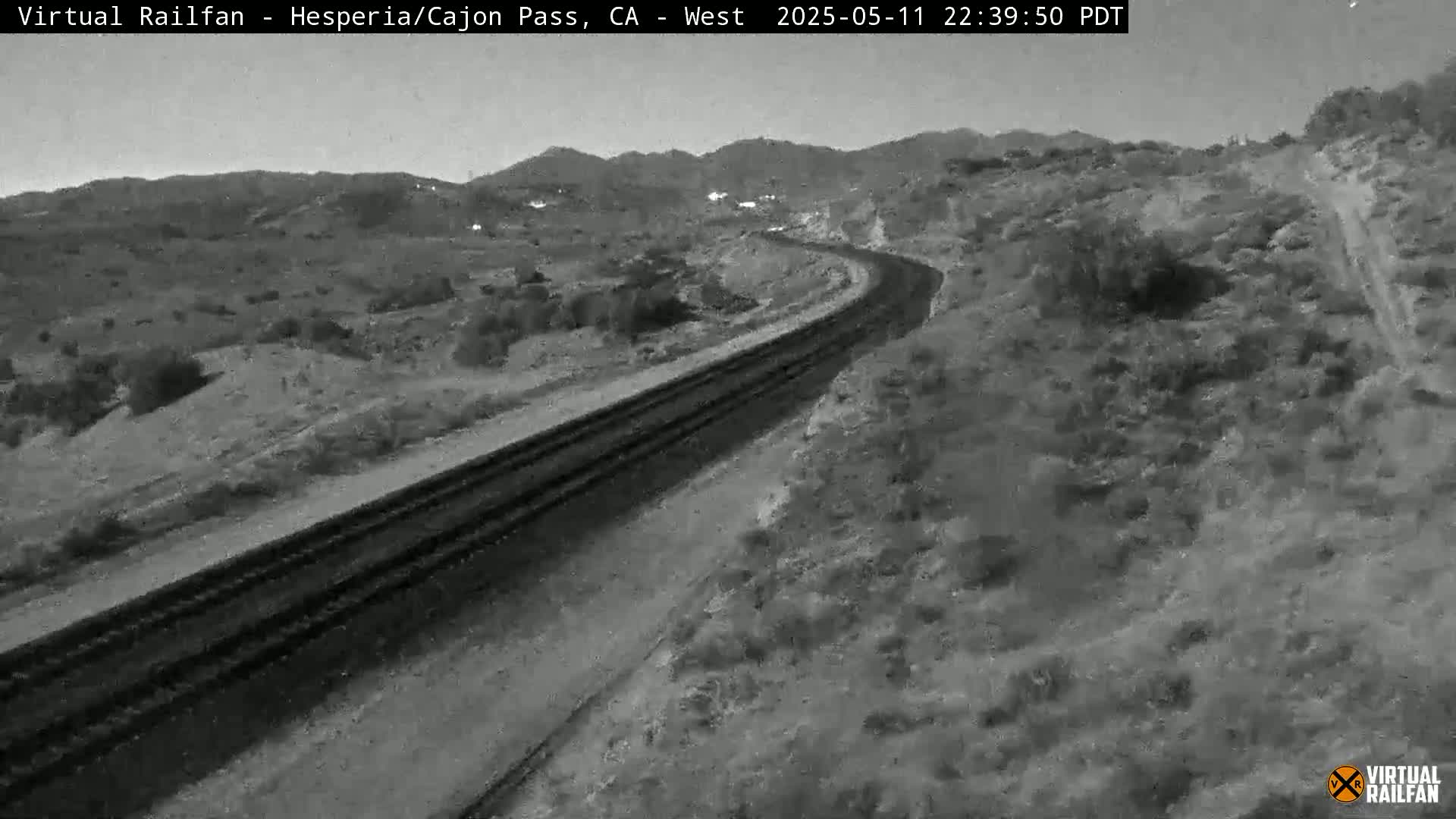 A nighttime, black-and-white view shows a curving train track winding through a hilly, desert-like landscape under a clear sky.