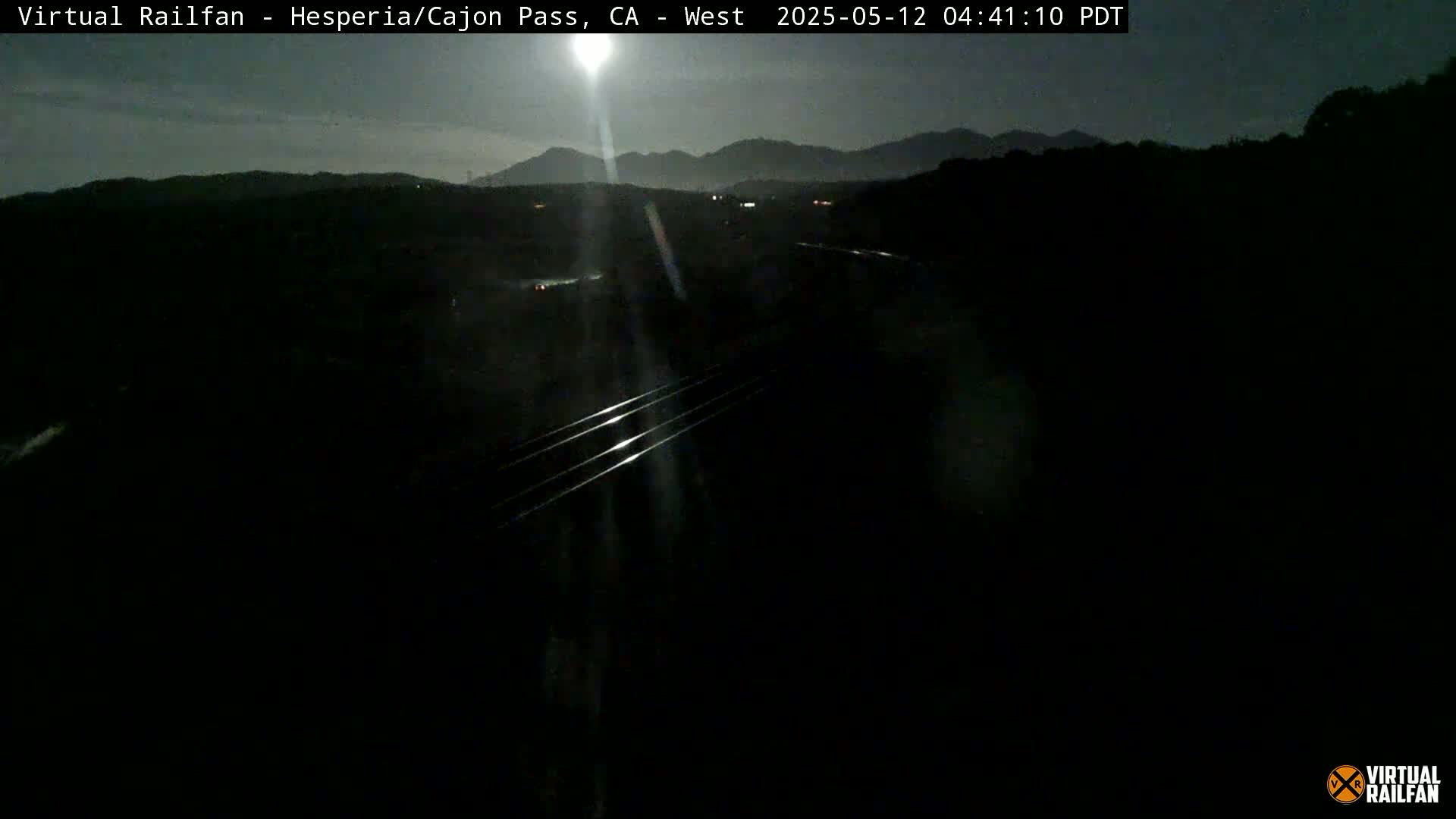 A nighttime view of a highway and railroad tracks, with mountains in the background under a mostly clear sky.