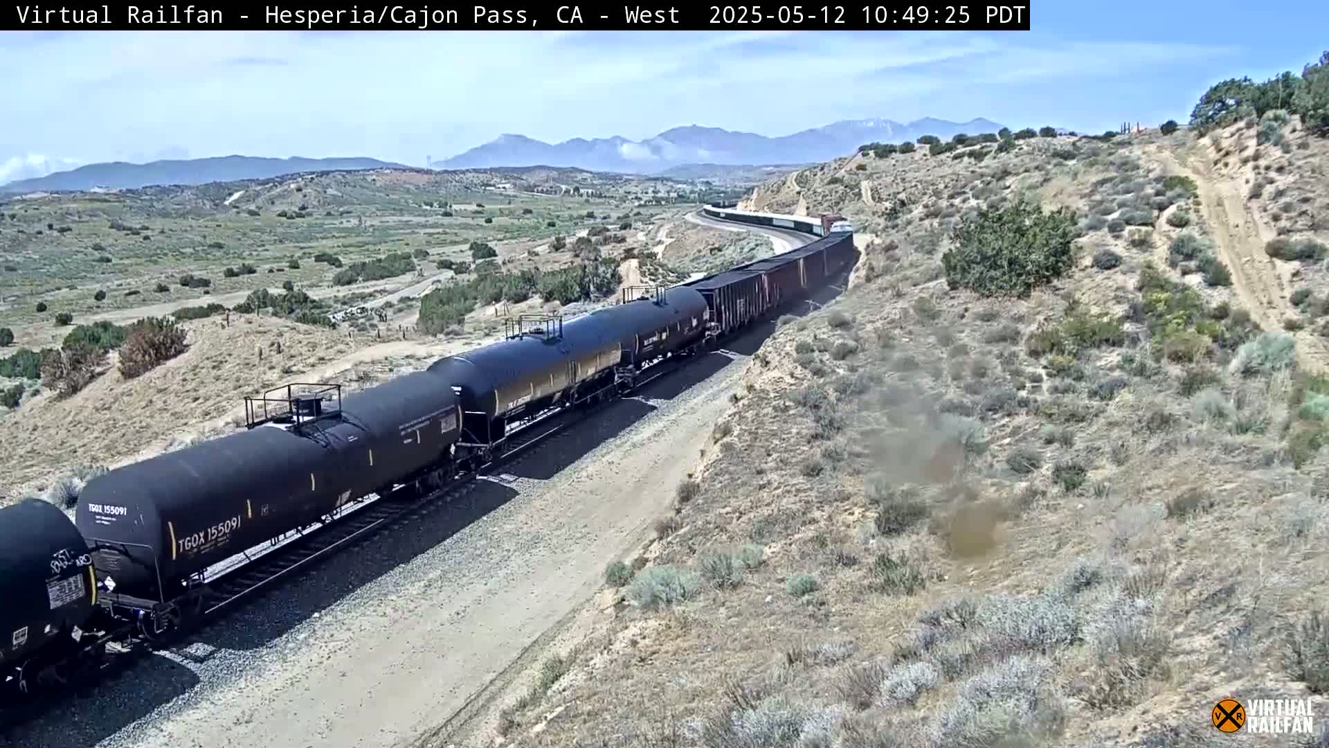 A long freight train of tanker cars and boxcars travels along a rail line through a dry, mountainous landscape under a clear sky.