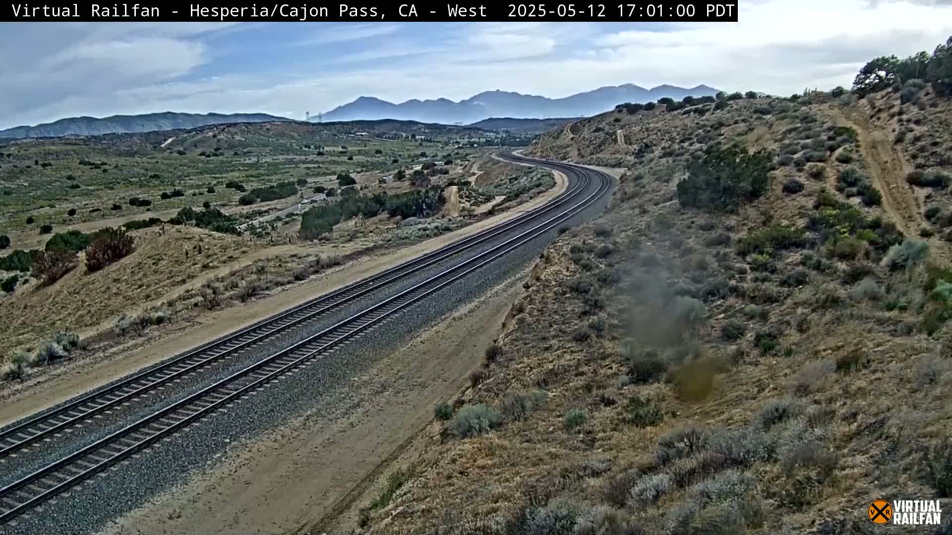 A mostly clear day reveals a view of double railroad tracks curving through a dry, mountainous landscape under a partly cloudy sky.