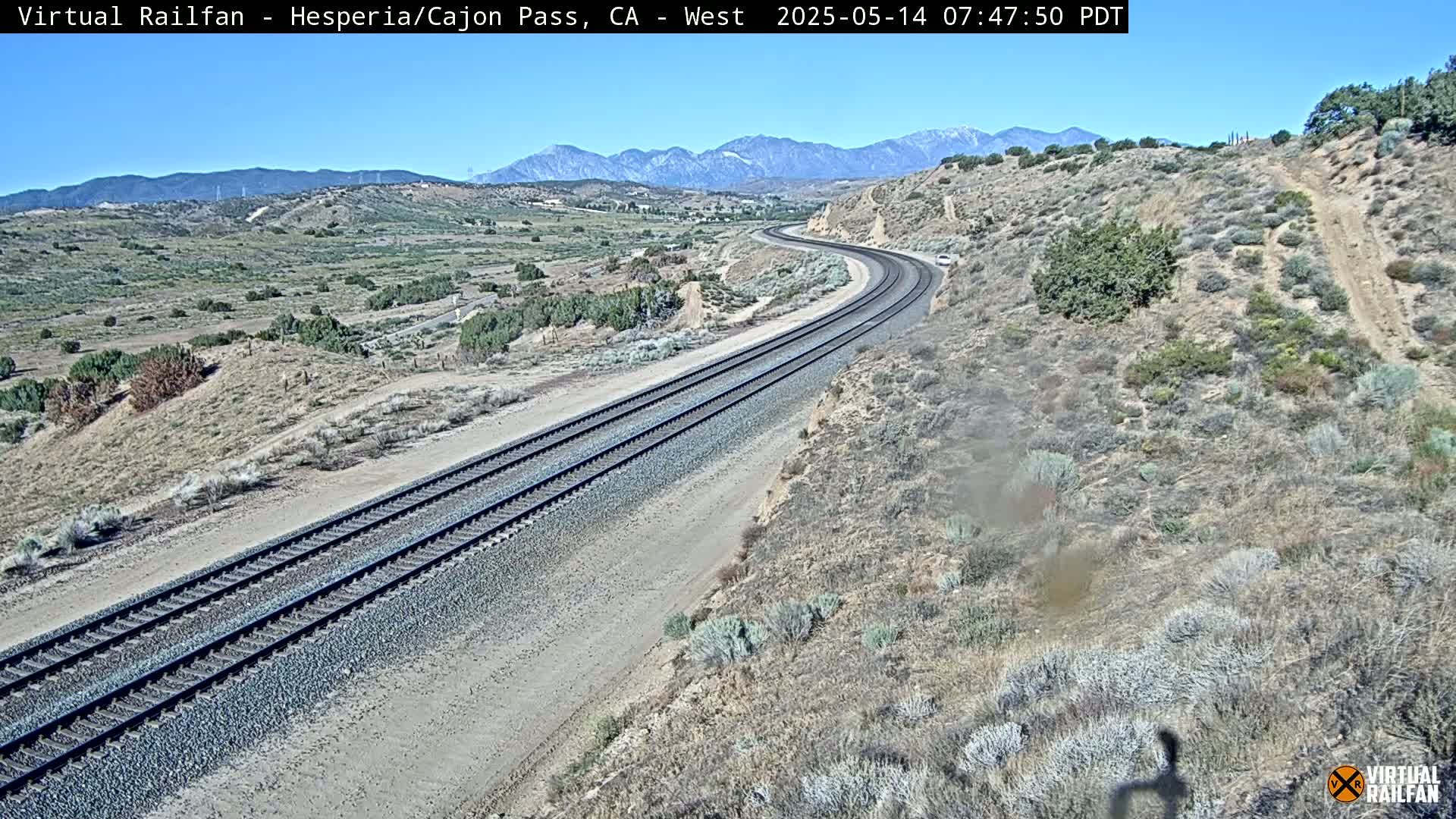 A pair of train tracks curves through a dry, scrub-covered landscape under a clear sky, with mountains visible in the distance.