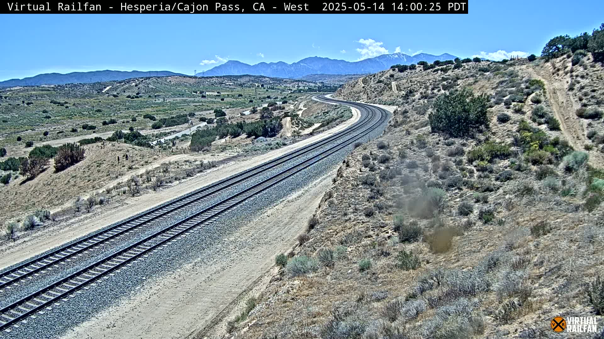 A high-angle view shows two parallel sets of train tracks curving gently through a dry, desert landscape under a clear, sunny sky with mountains in the background.