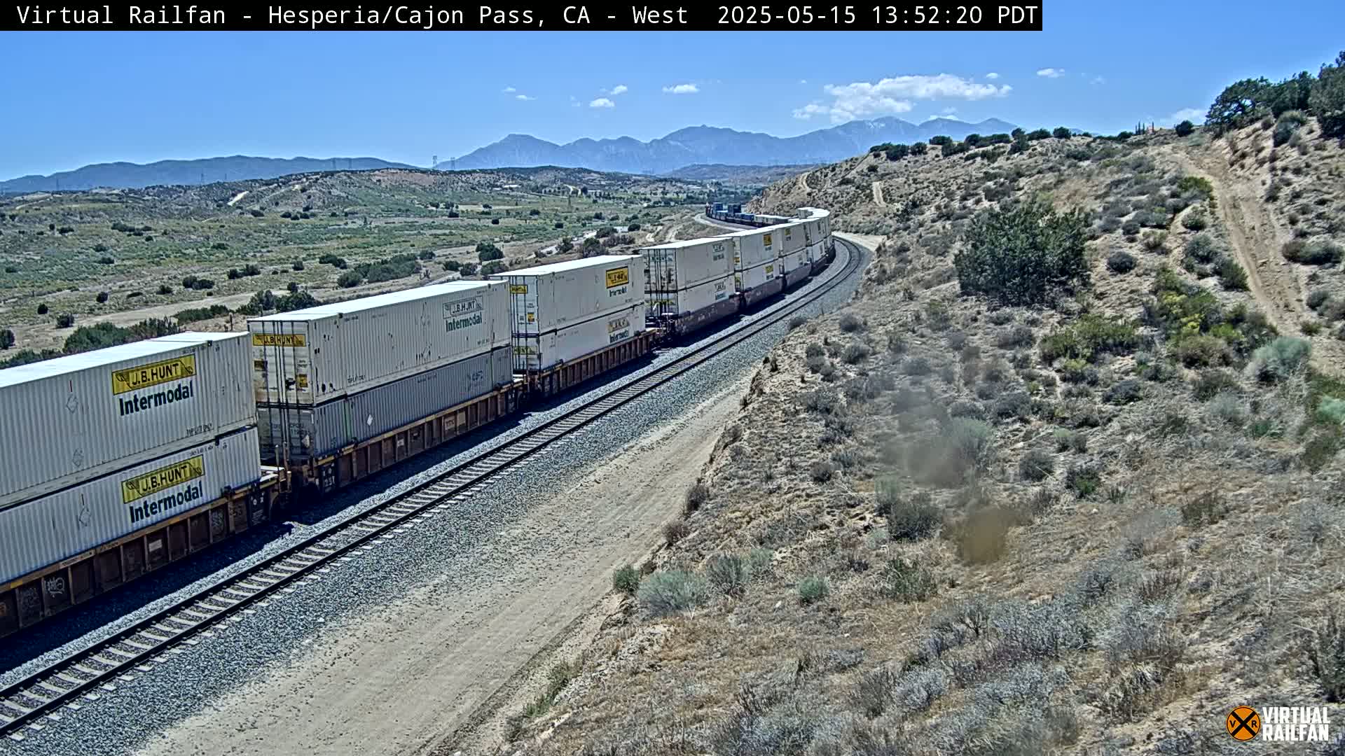 A long freight train carrying multiple shipping containers moves along a railway track through a dry, desert-like landscape under a clear blue sky.