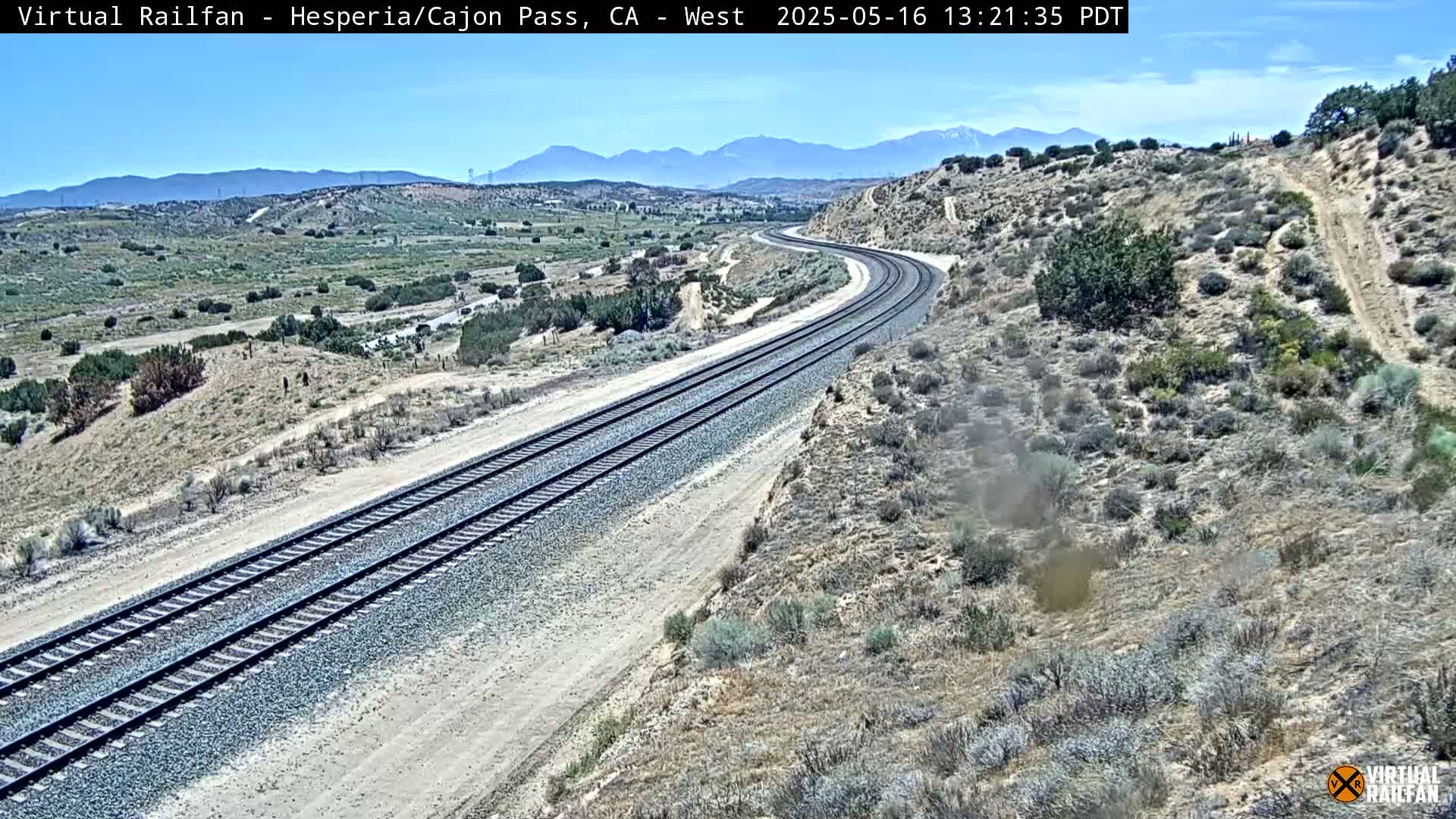A long stretch of railroad tracks curves gently through a dry, desert-like landscape under a clear blue sky.