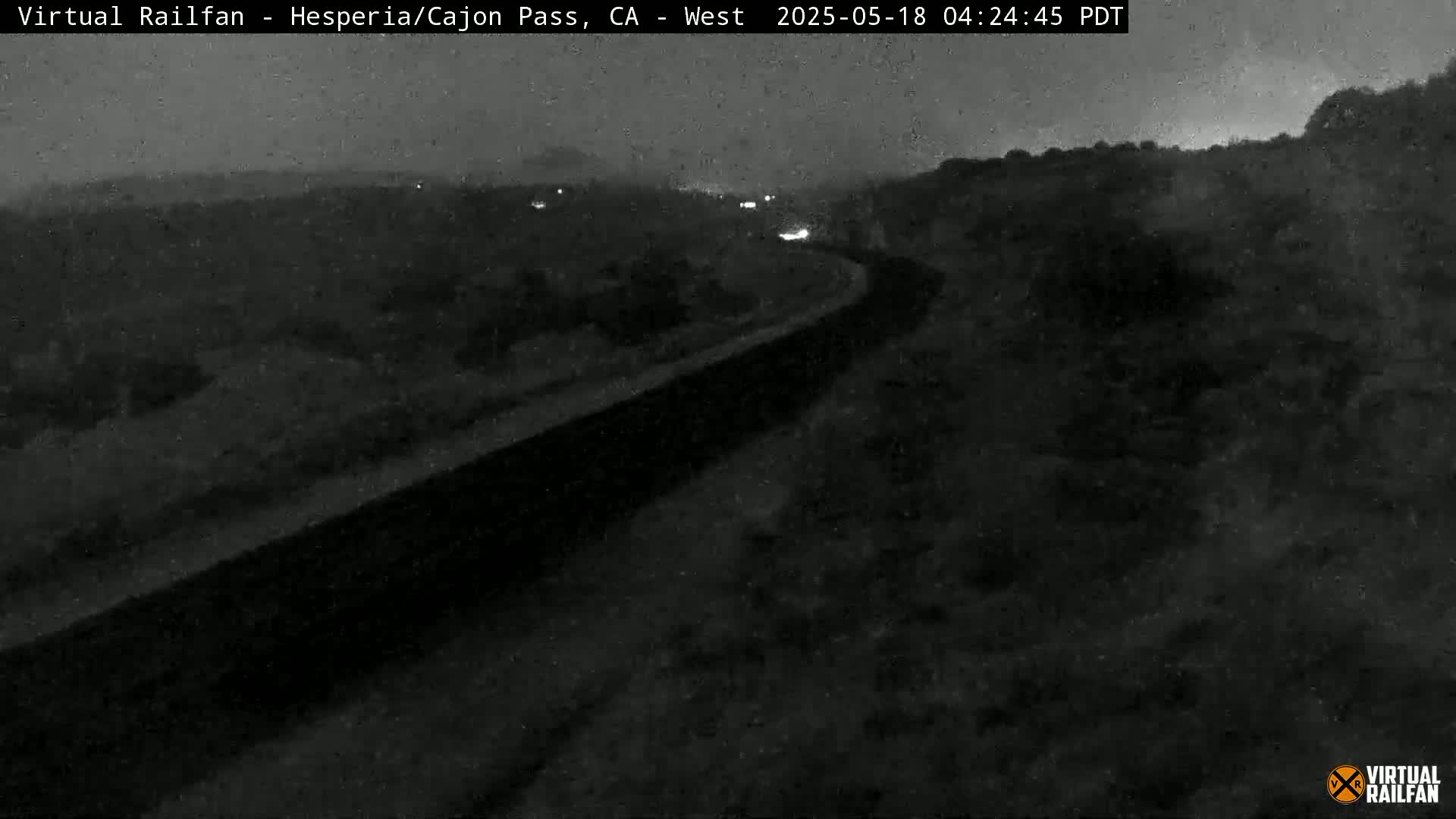 A nighttime, low-light view shows a dark roadway curving through a sparsely vegetated landscape.