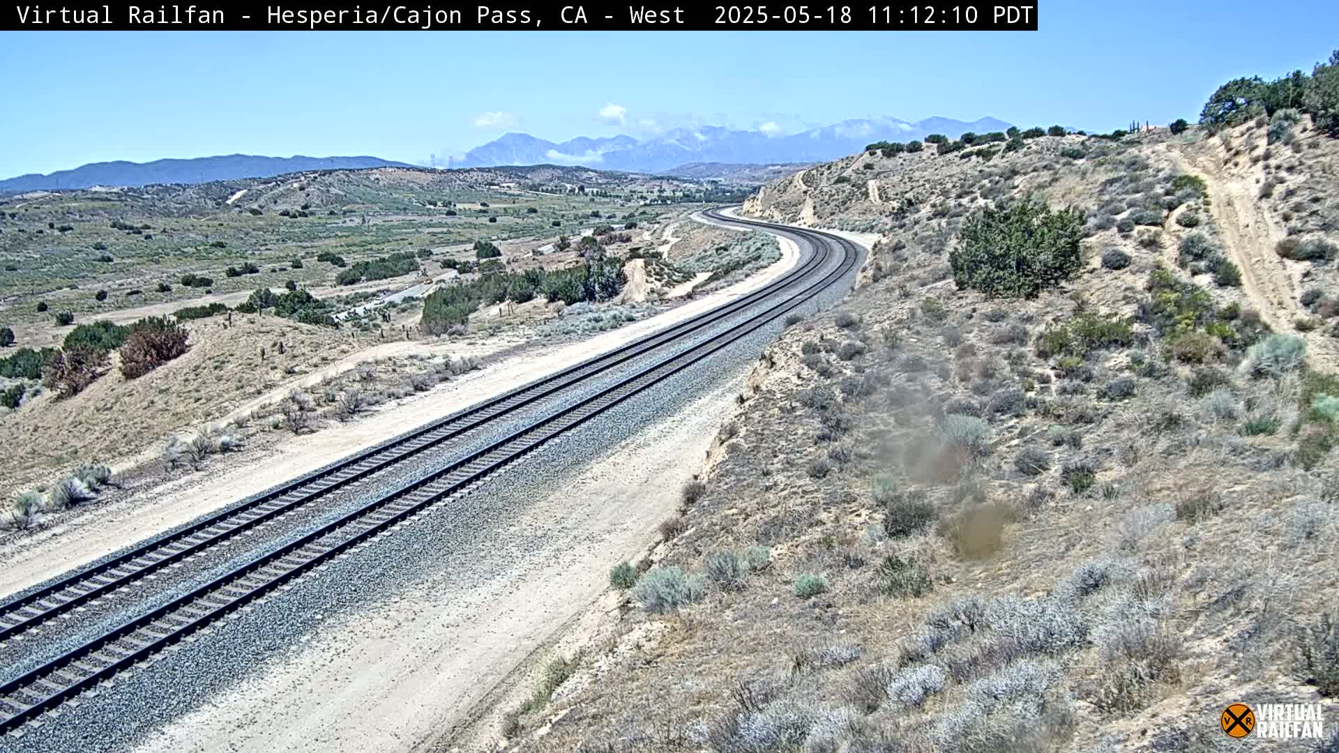 A high-angle view shows a curving train track traversing a dry, desert-like landscape under a clear sky with distant mountains.