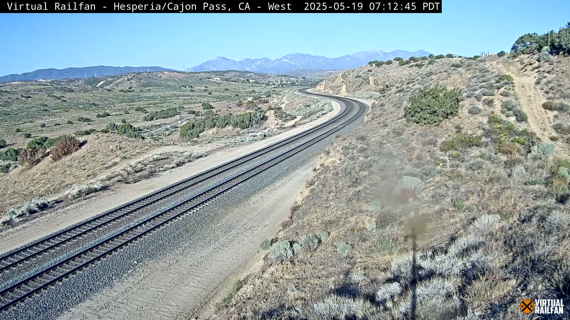A high-angle view of a curving railway track traversing a dry, desert-like landscape under a clear, sunny sky with mountains in the background.