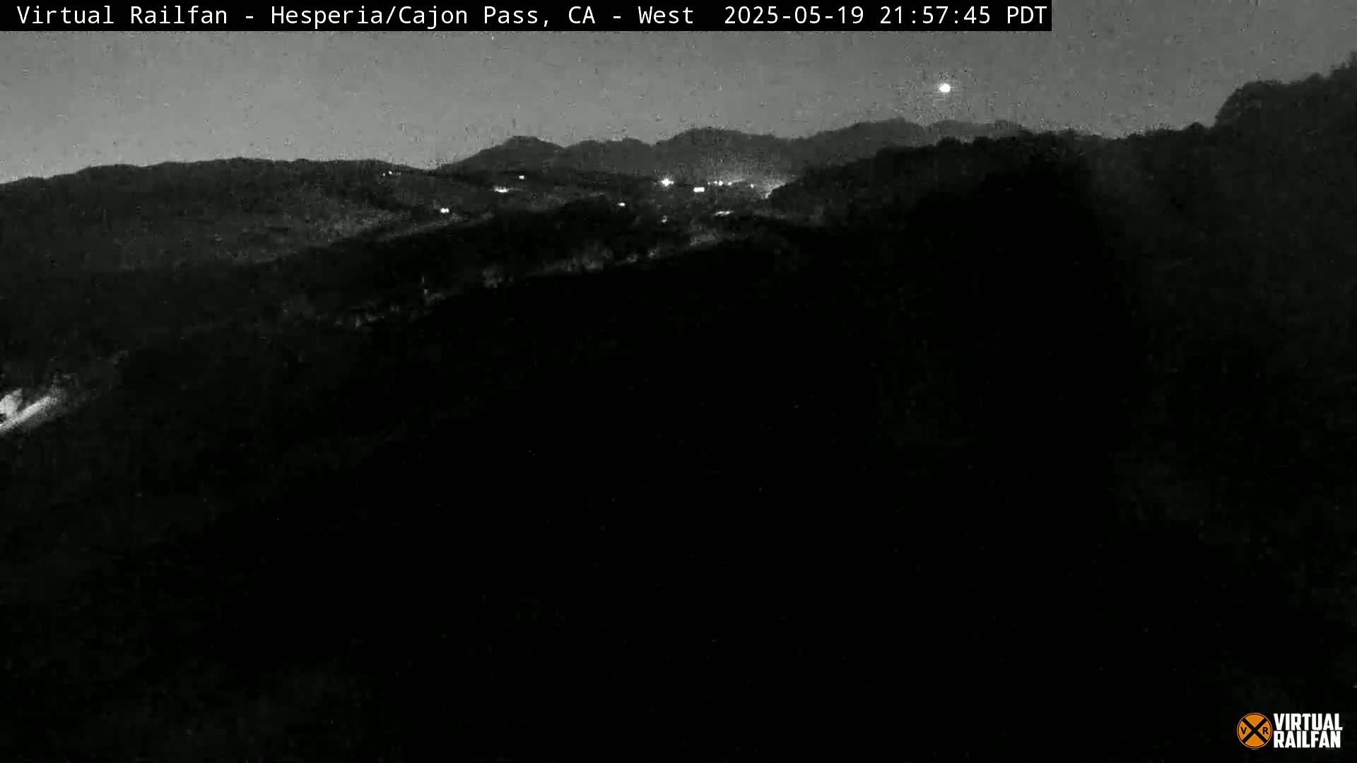 A nighttime, low-light view shows dark hills and a valley with a few distant lights under a clear, dark sky.