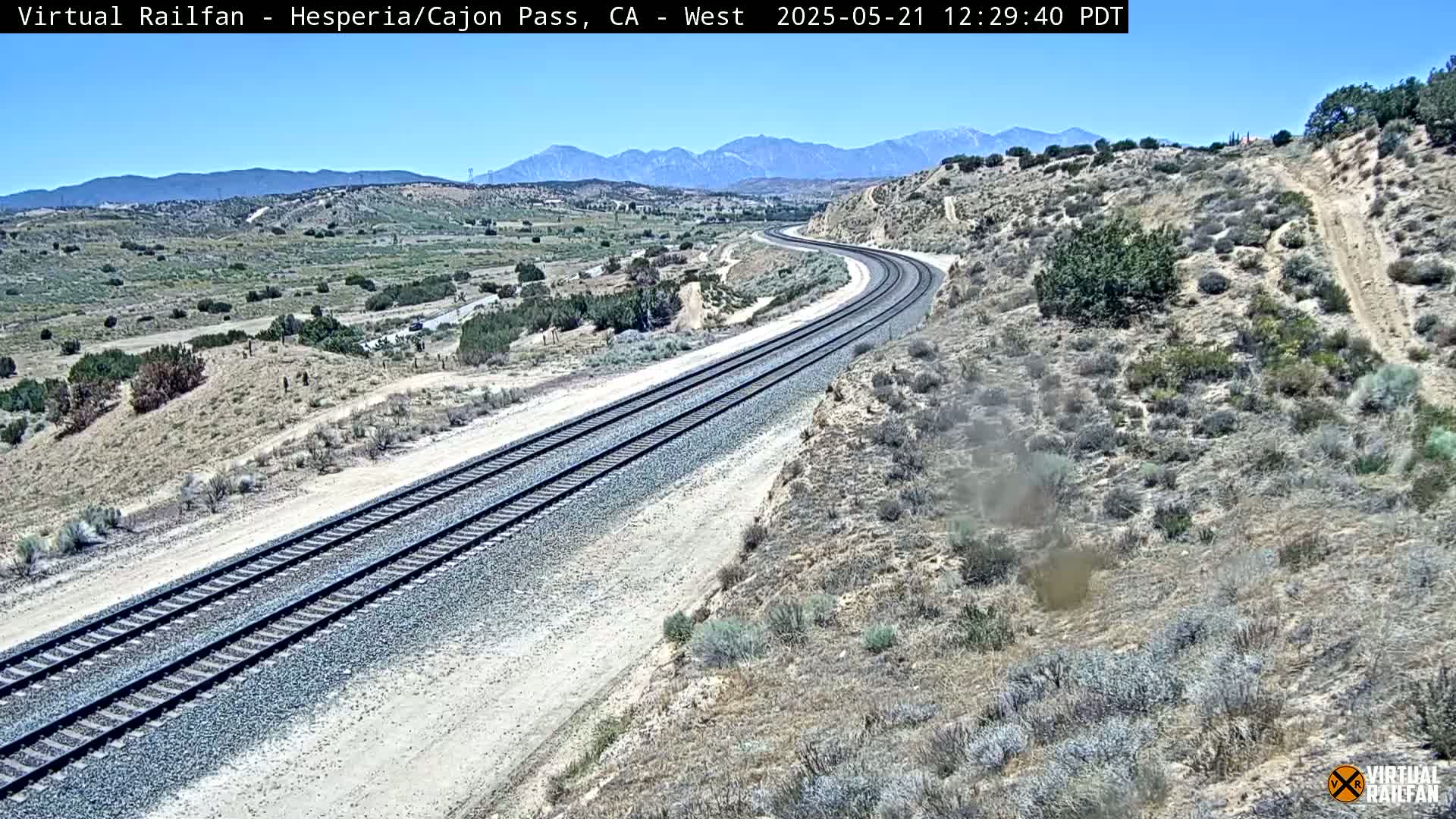 A long, curving train track stretches across a dry, desert landscape under a clear blue sky, with mountains visible in the distance.