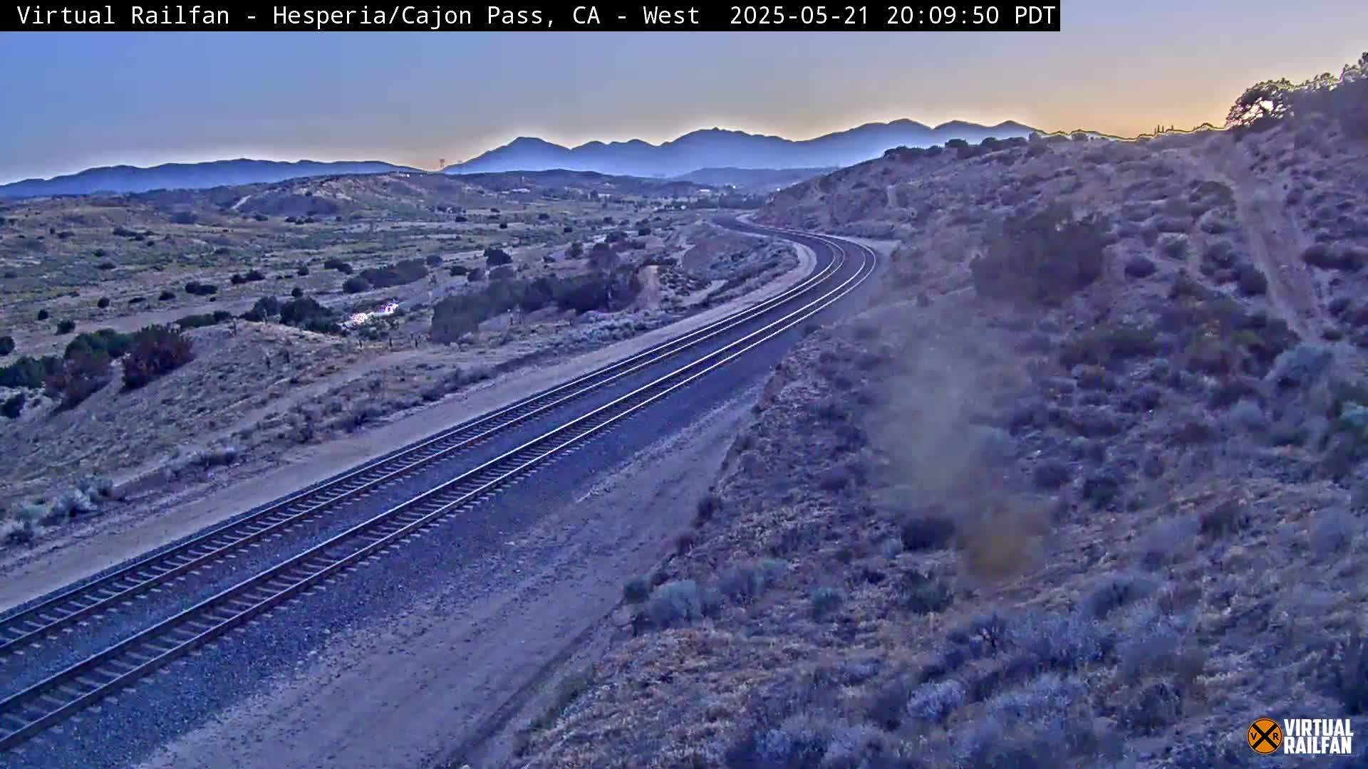 A high-angle, long shot shows a pair of train tracks curving through a desert landscape at dusk under a clear sky.