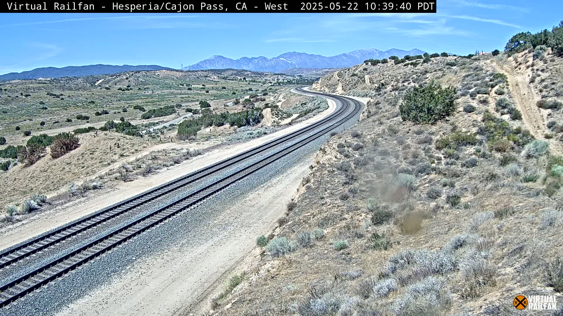 A high-angle view of a curving train track in a dry, desert-like landscape under a clear sky, with mountains in the background.