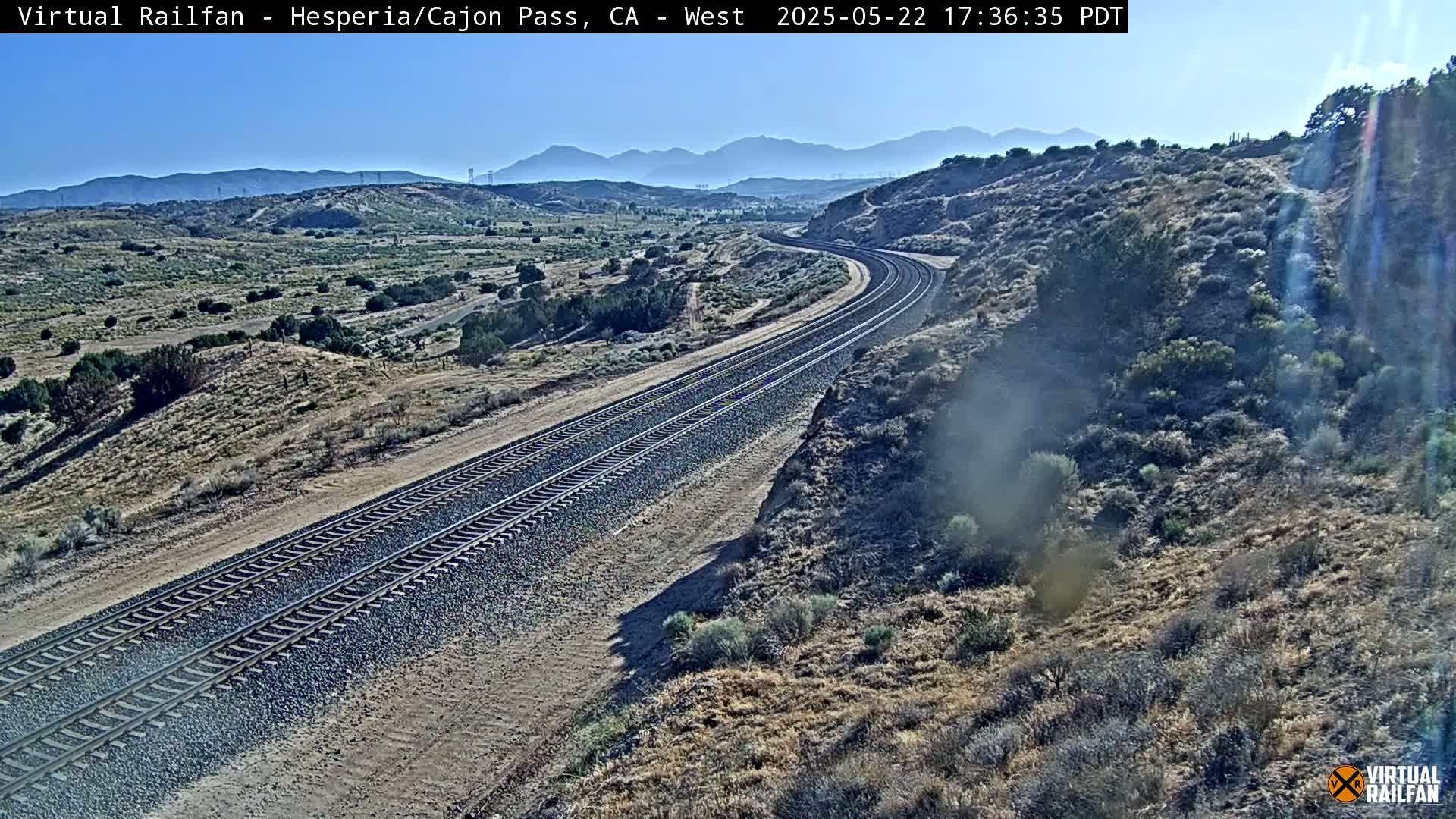 A long stretch of train tracks curves through a dry, mountainous landscape under a clear, sunny sky.