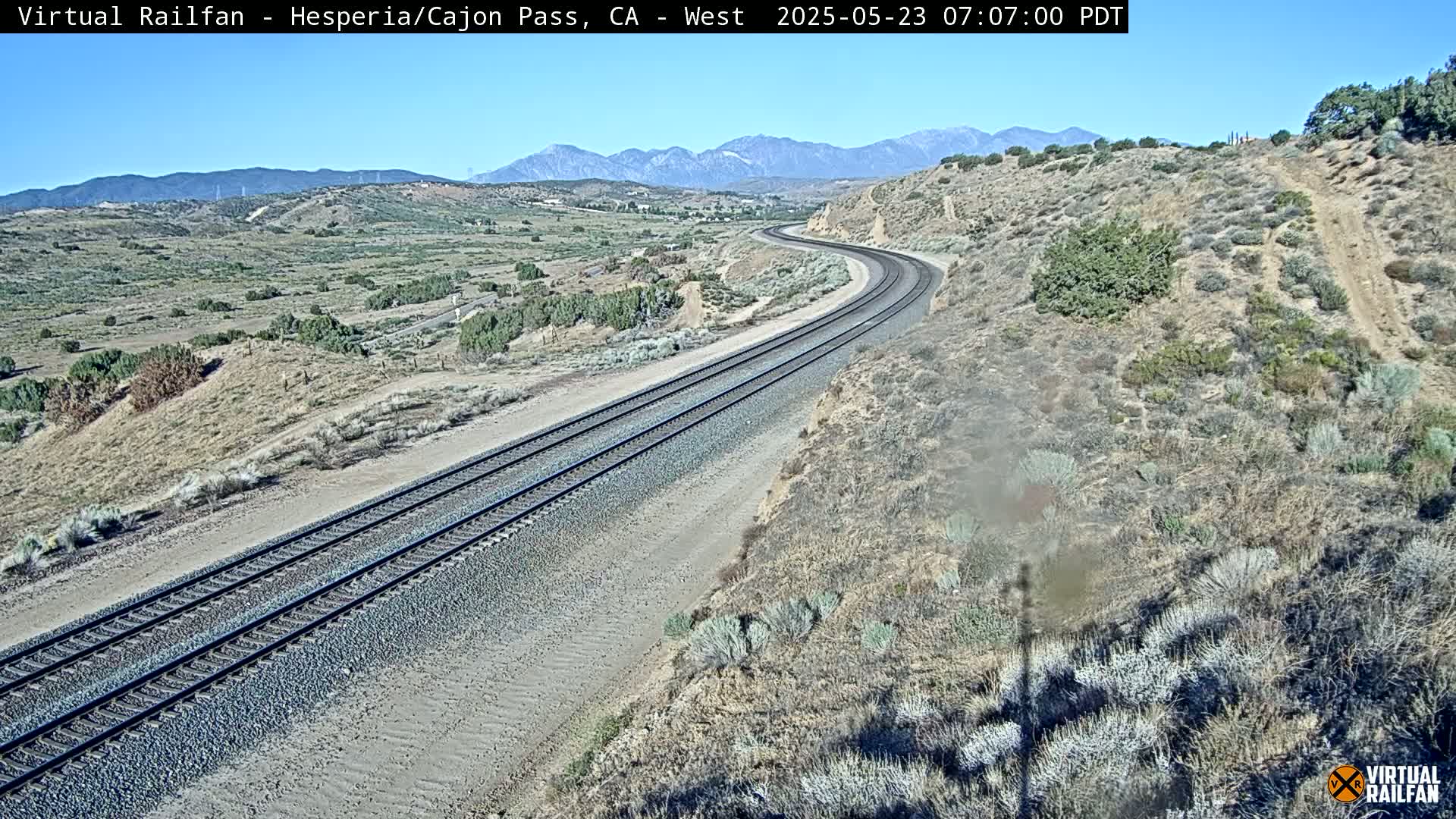 A curving railroad track winds through a dry, scrub-covered landscape under a clear blue sky, with mountains visible in the distance.